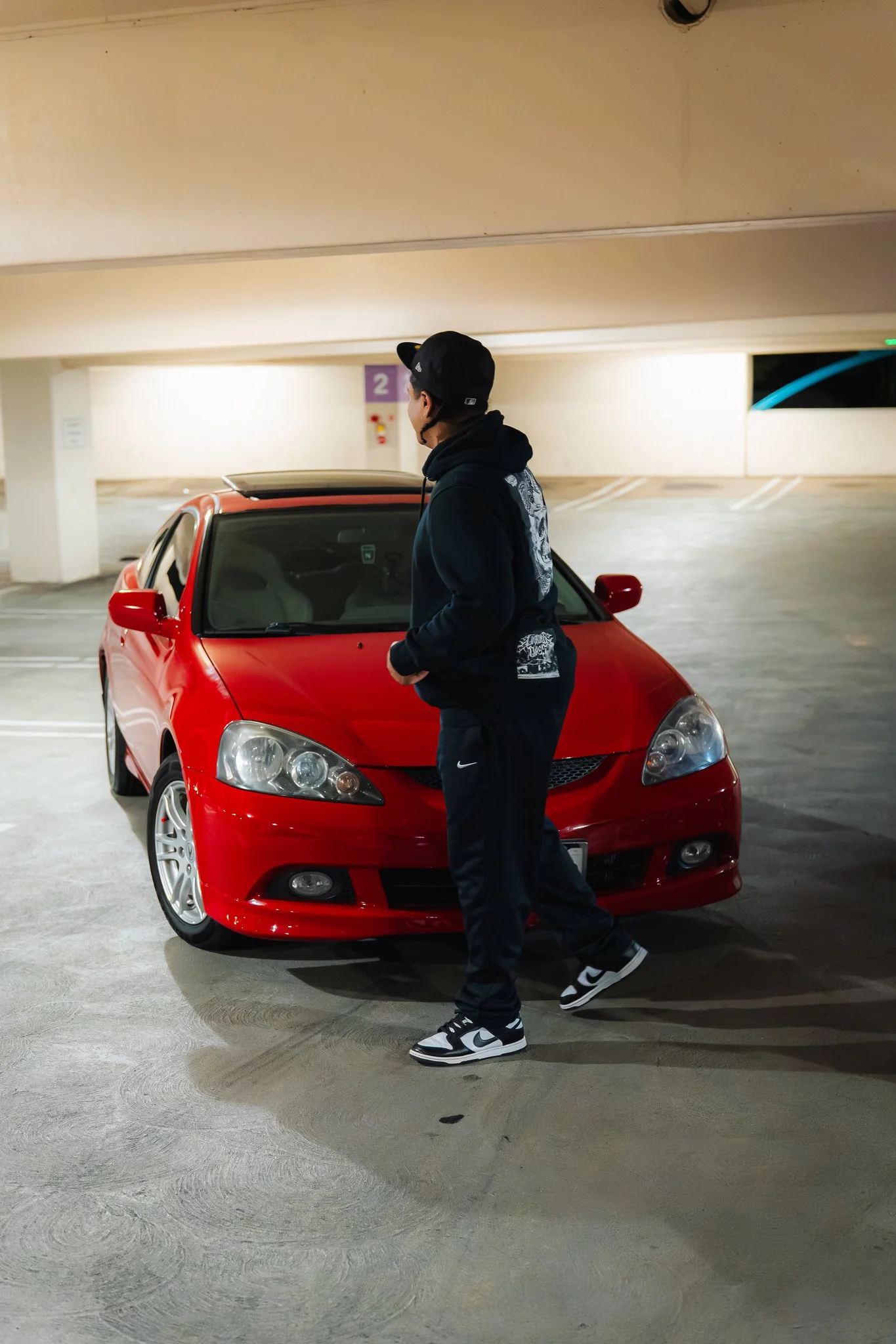 A man in black Nike sneakers and black pants, wearing a black hoodie with a graphic design on the back, and a black baseball cap, is walking past a red car in an indoor parking garage.