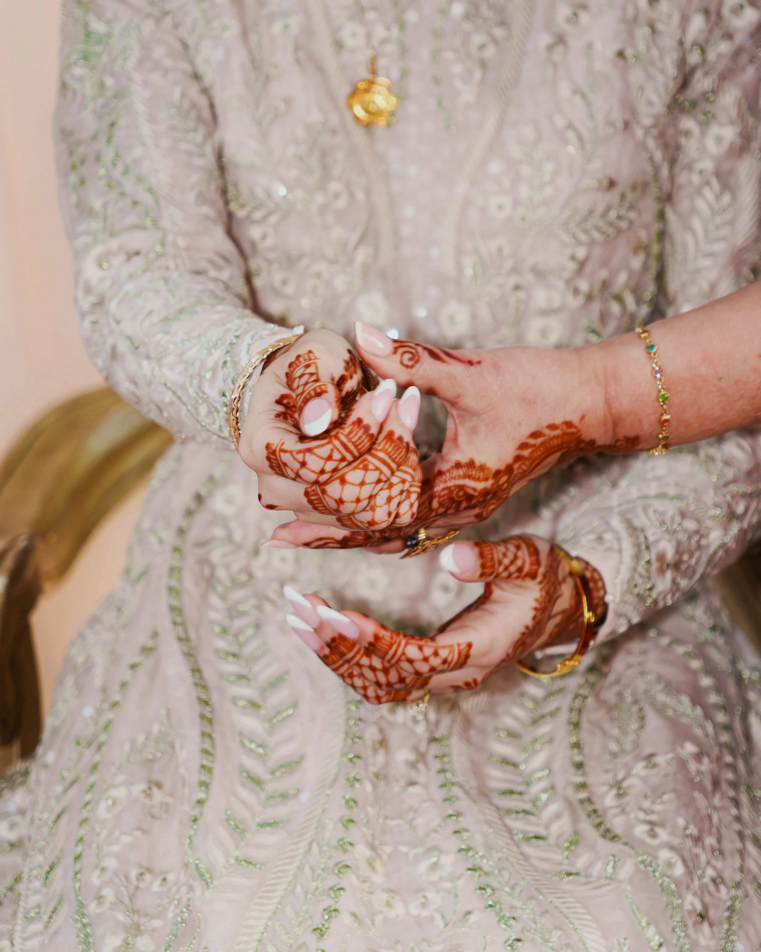 Close-up of a person wearing traditional Indian clothing and jewelry, with detailed henna designs on their hands, adjusting a ring on their finger. The background features an ornate, embroidered fabric with silver and green embellishments.