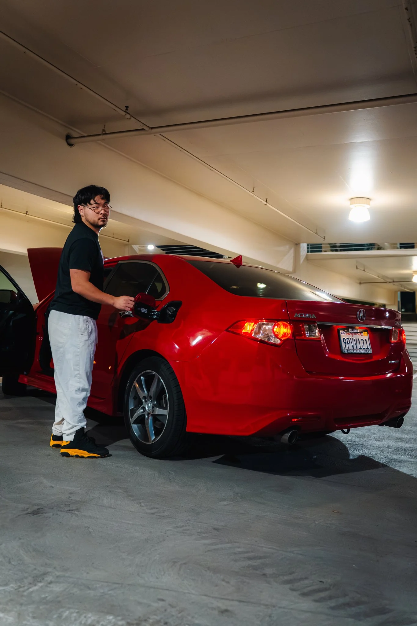 A young man with glasses and black hair, dressed in a black t-shirt, white pants, and black and yellow shoes, stands next to a red car in an indoor parking garage. The man appears to be opening the driver's side door of the car.