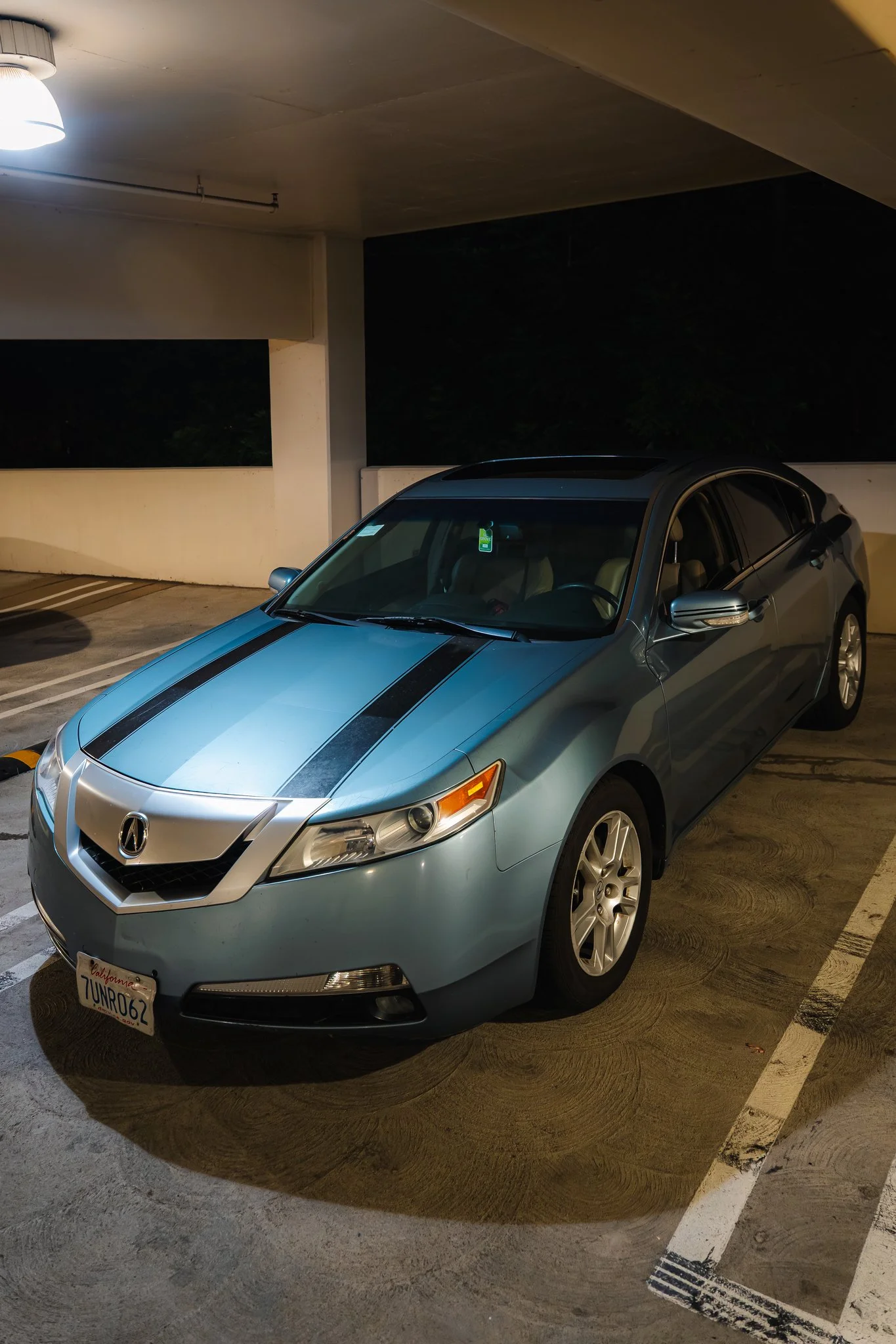 A silver Acura sedan parked in a dimly lit parking garage at night. The car has a sunroof, and a California license plate reading 7UNR062. The parking lot is mostly empty with marked parking spaces and a white concrete barrier in the background.