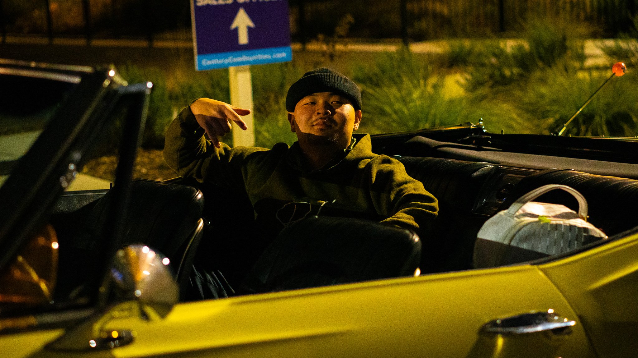 A man sitting in a yellow convertible car at night making a peace sign with his right hand, wearing a black beanie and a green jacket, with a street sign visible in the background.