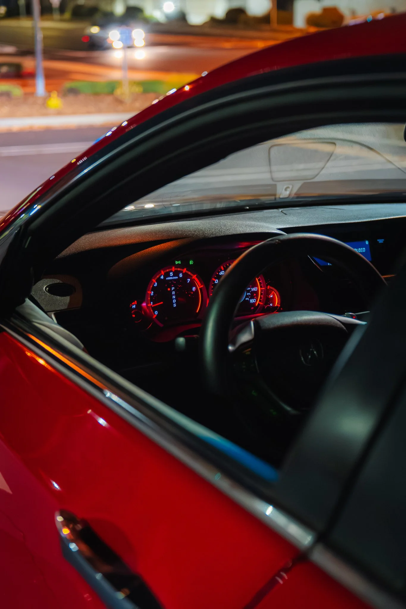 Close-up view from inside a red car at night, with the dashboard illuminated and the blurred city street visible through the windshield.