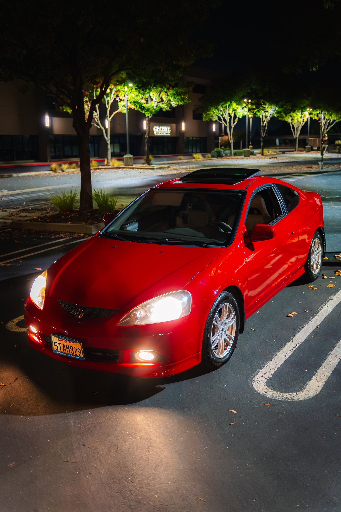 A red compact car parked in a lot at night with trees and building in the background.
