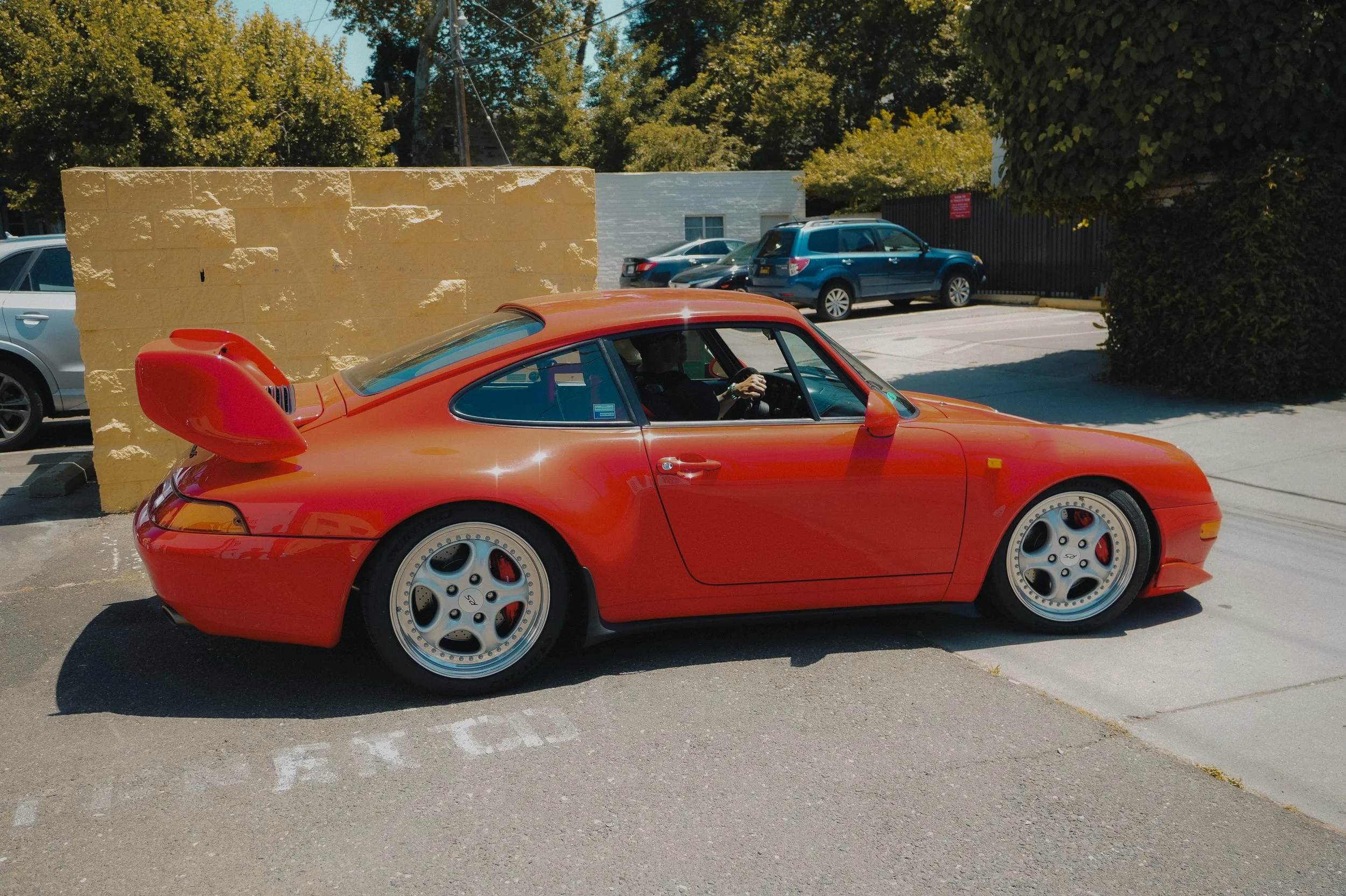 A bright red sports car with a large rear spoiler parked in a lot with other vehicles and a yellow brick wall in the background.