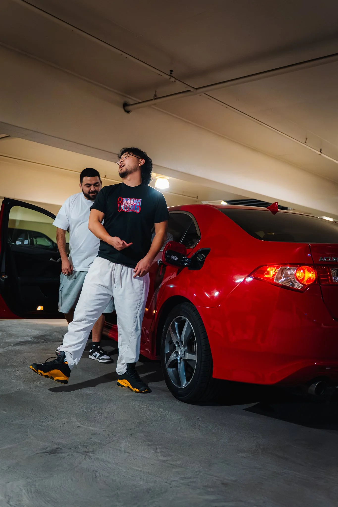 Two young men standing next to a red car in an underground parking garage, one of them wearing black sneakers and yellow accents, the other in white and black sneakers, both casually dressed.