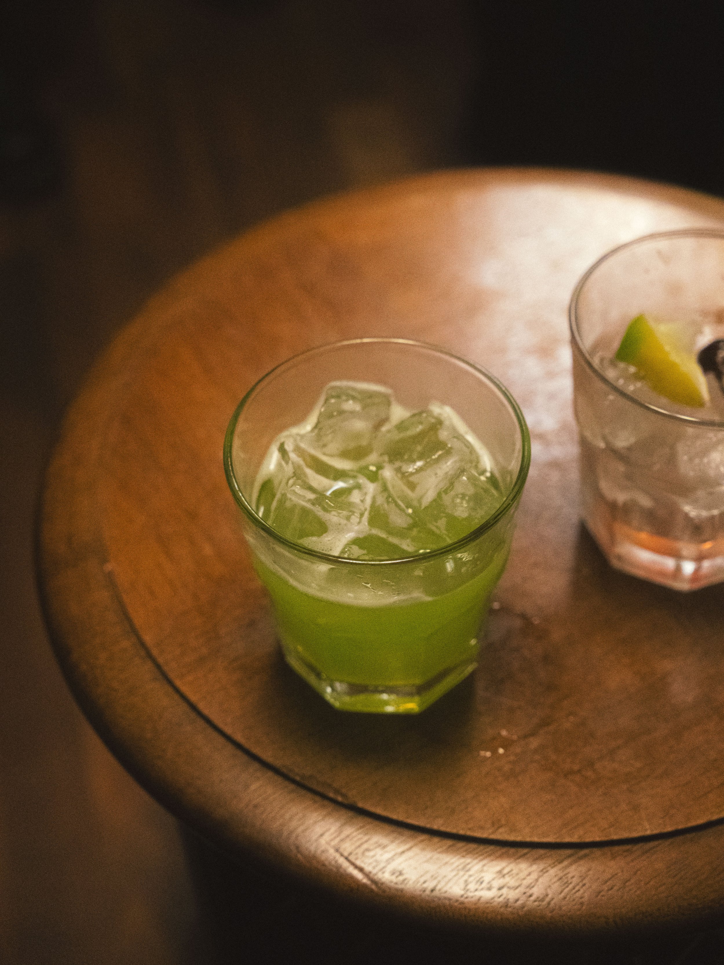 A glass of green-colored beverage with ice cubes and some lime slices on top, next to a glass with clear liquid and lime wedge, on a wooden table.