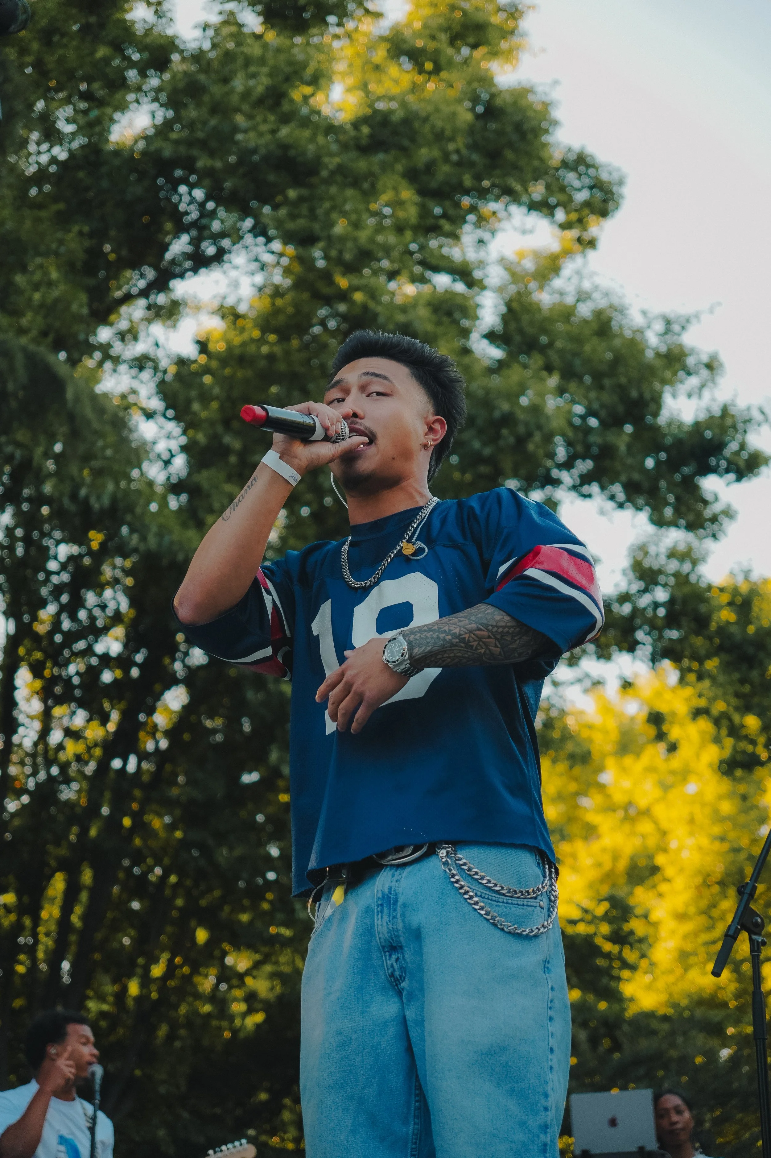 A young man with dark hair holding a microphone in an outdoor setting, surrounded by trees and other people.