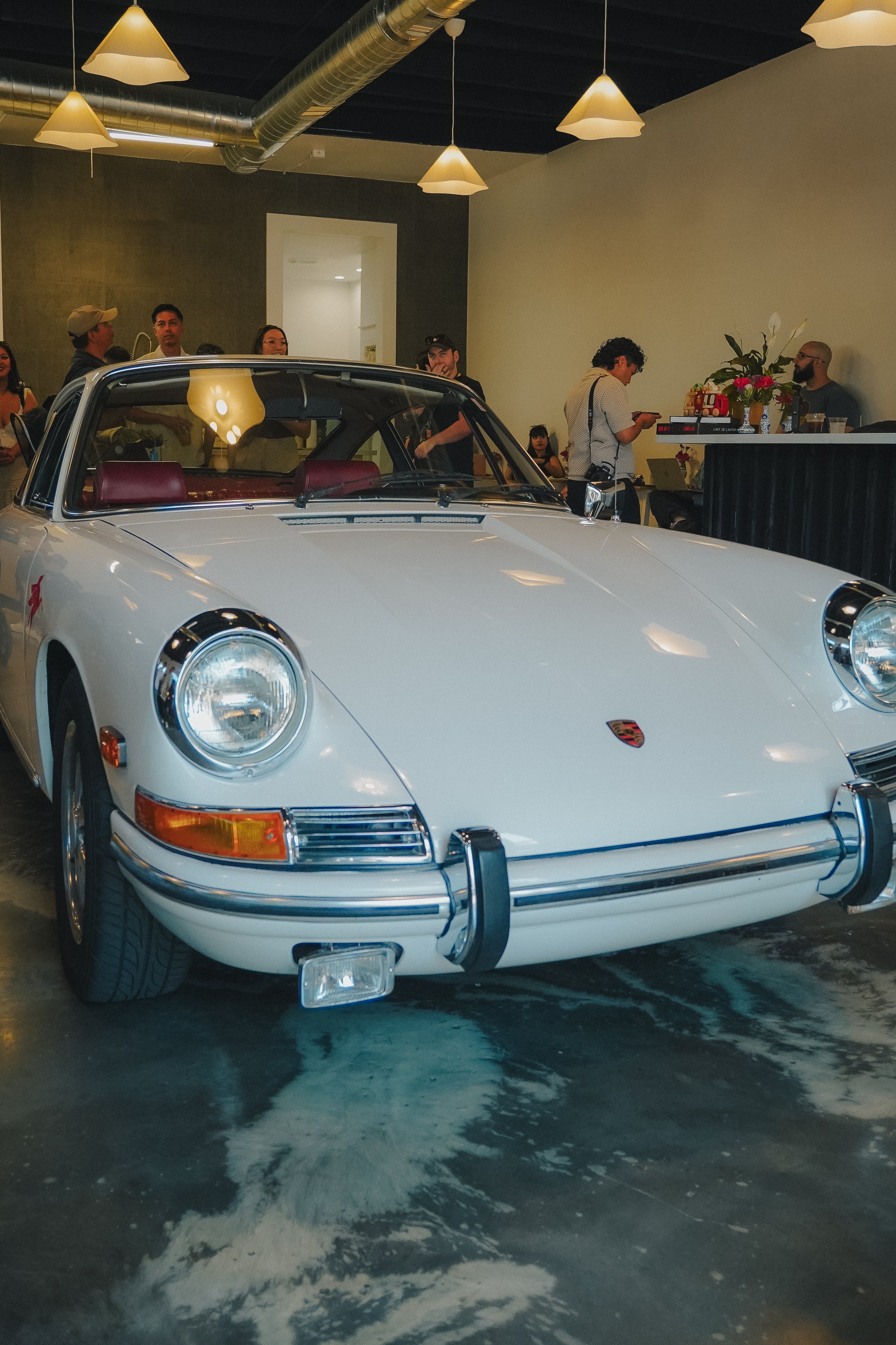 A vintage white Porsche 911 parked indoors with a group of people gathered around and talking. The background features a black wall with hanging light fixtures and an indoor plant with flowers on a counter.