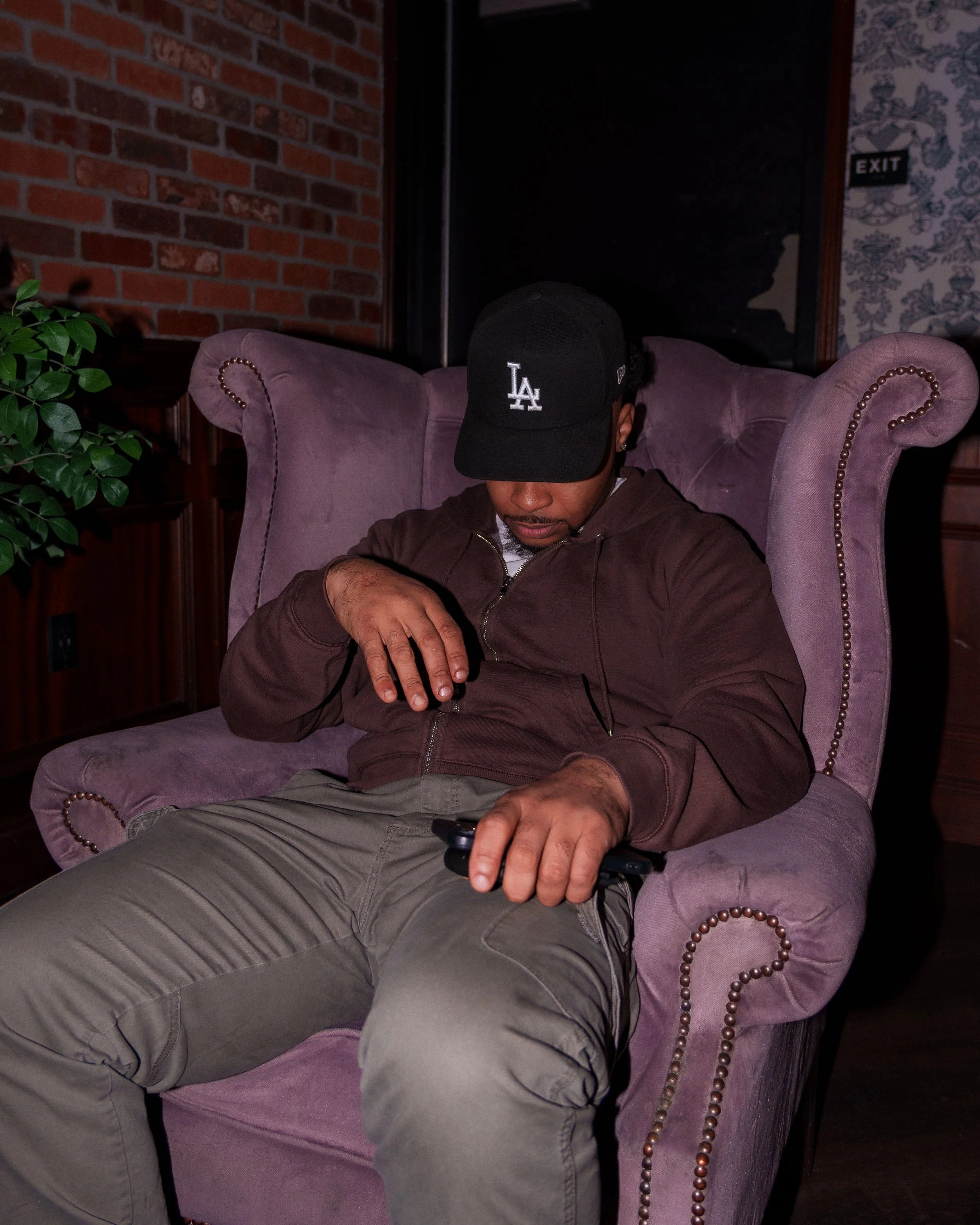 A person sitting in a purple armchair in a dimly lit room, wearing a black cap with 'LA' logo, brown jacket, beige pants, holding a game controller, in front of a brick wall with a plant nearby.