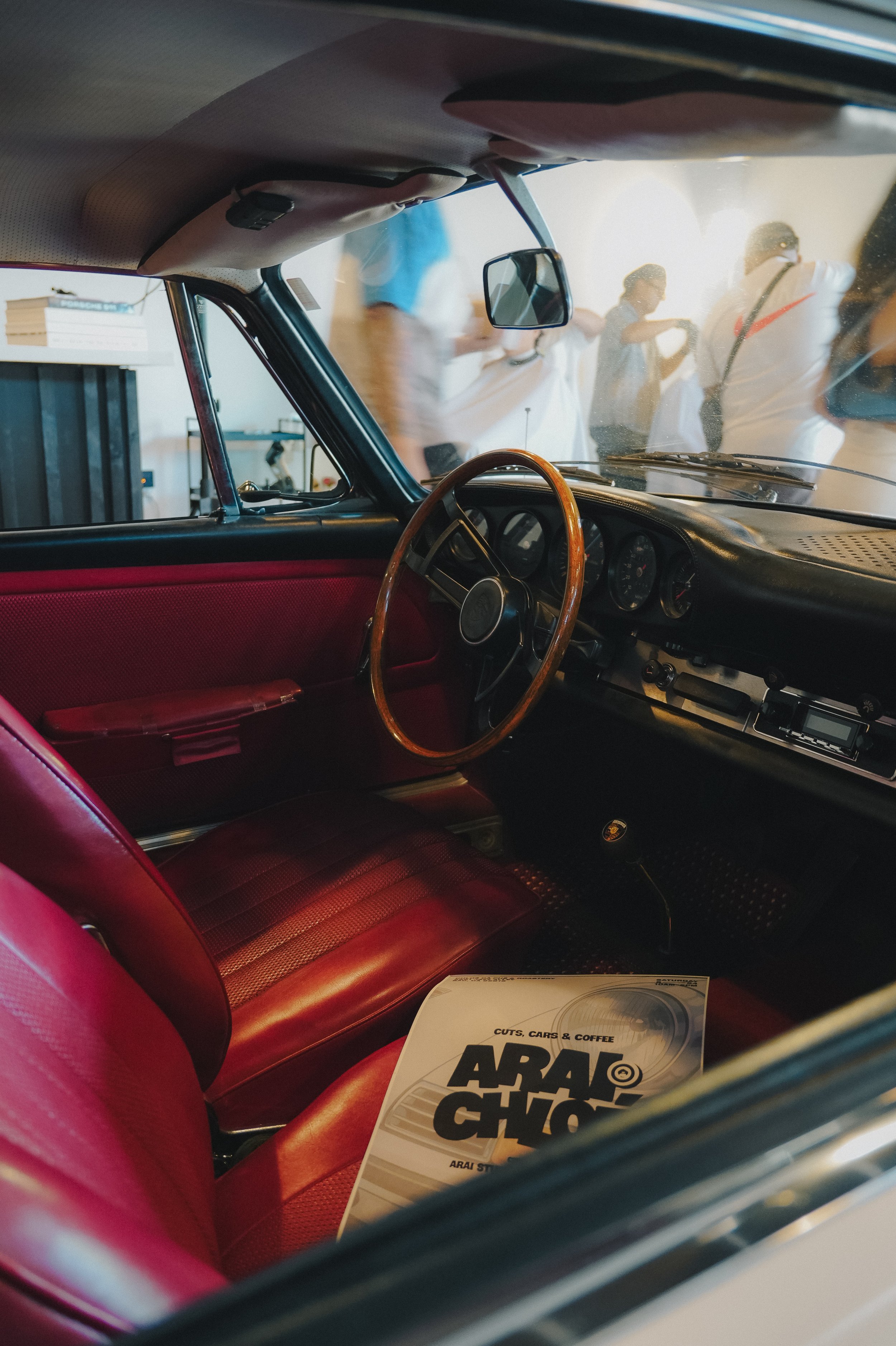 Inside view of a car with a red leather seat and a wooden steering wheel, showing some people outside through the windshield and a brochure on the dashboard titled "ARAI Auto Chop".