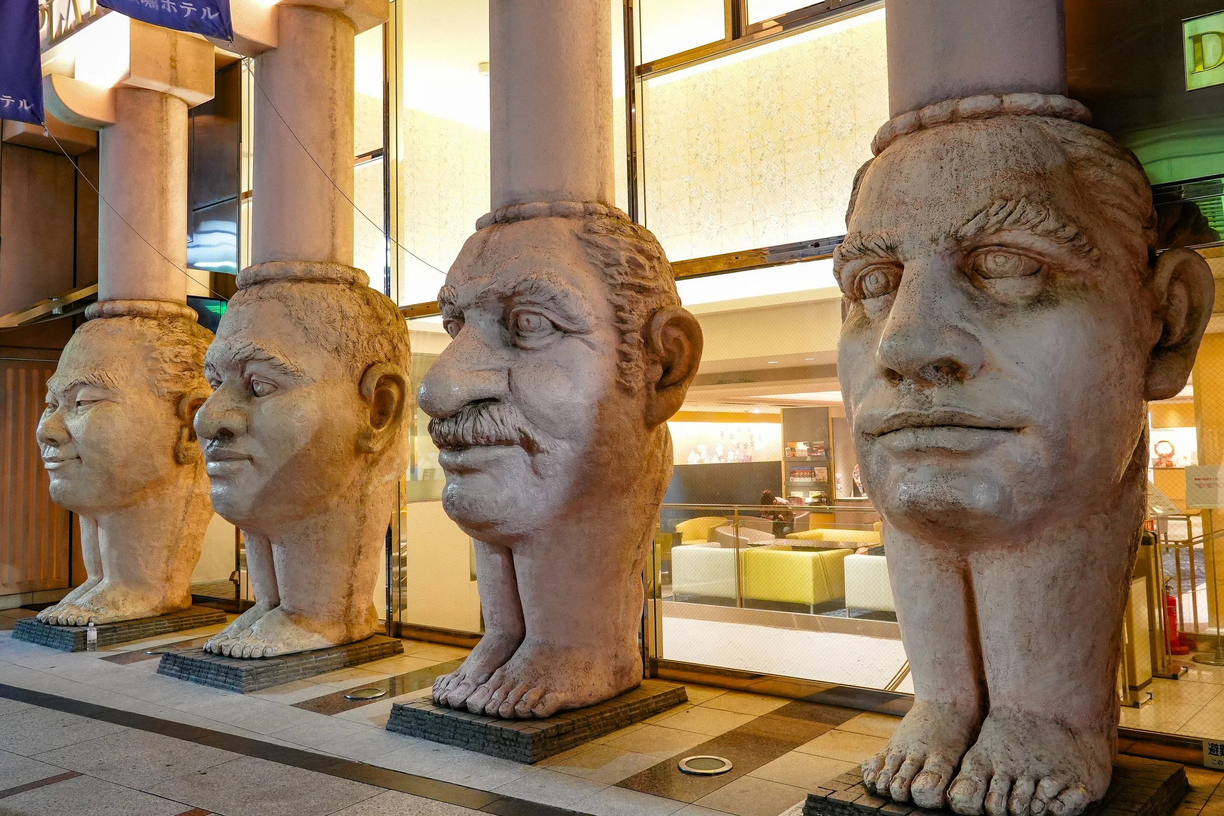 Four large stone statues of human heads with dog-like bodies and feet, displayed outdoors in front of a building.