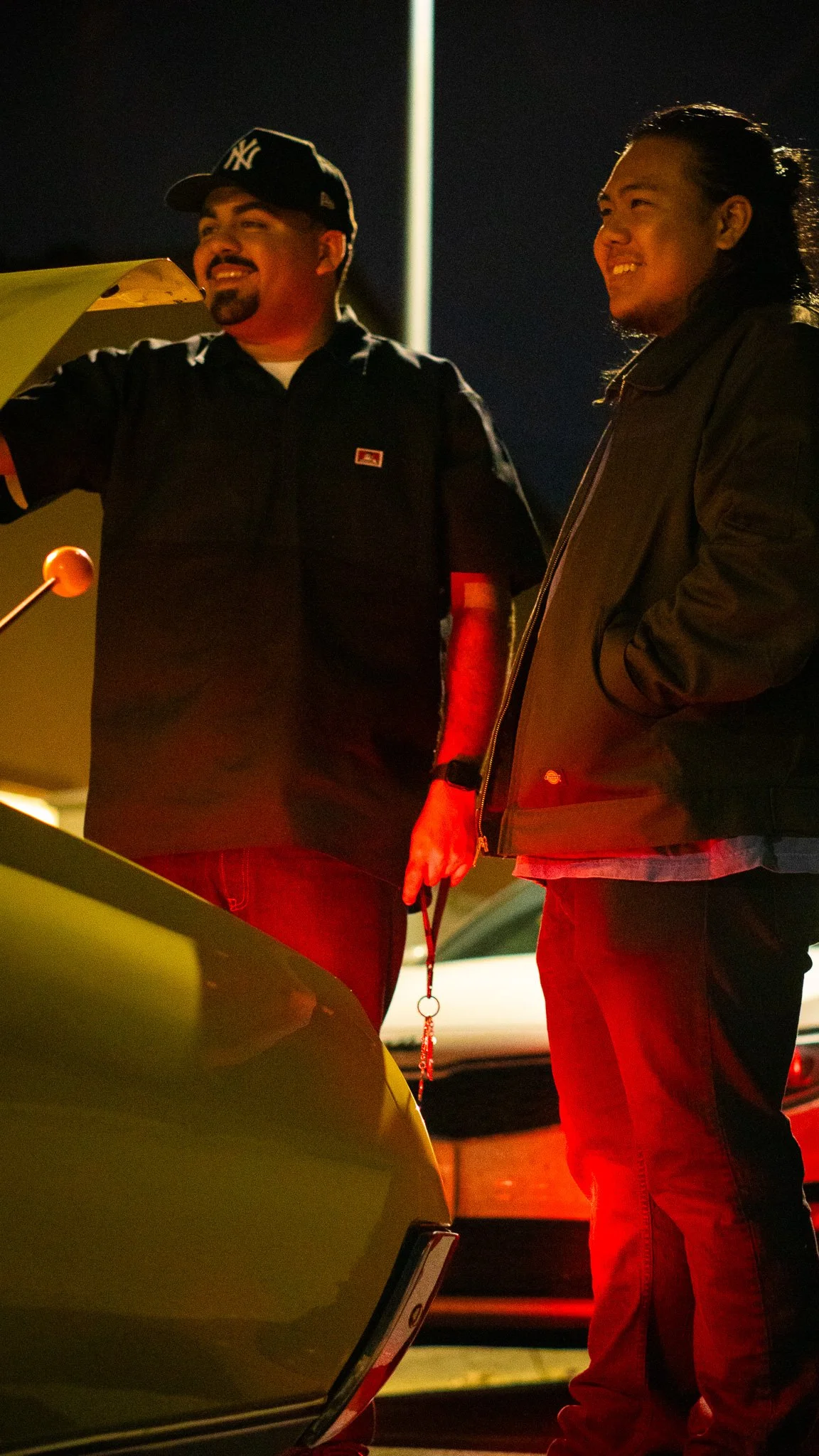 Two men standing at night near a yellow car, smiling and talking. One man wears a New York Yankees cap and a dark button-up shirt, the other a dark jacket and red pants.