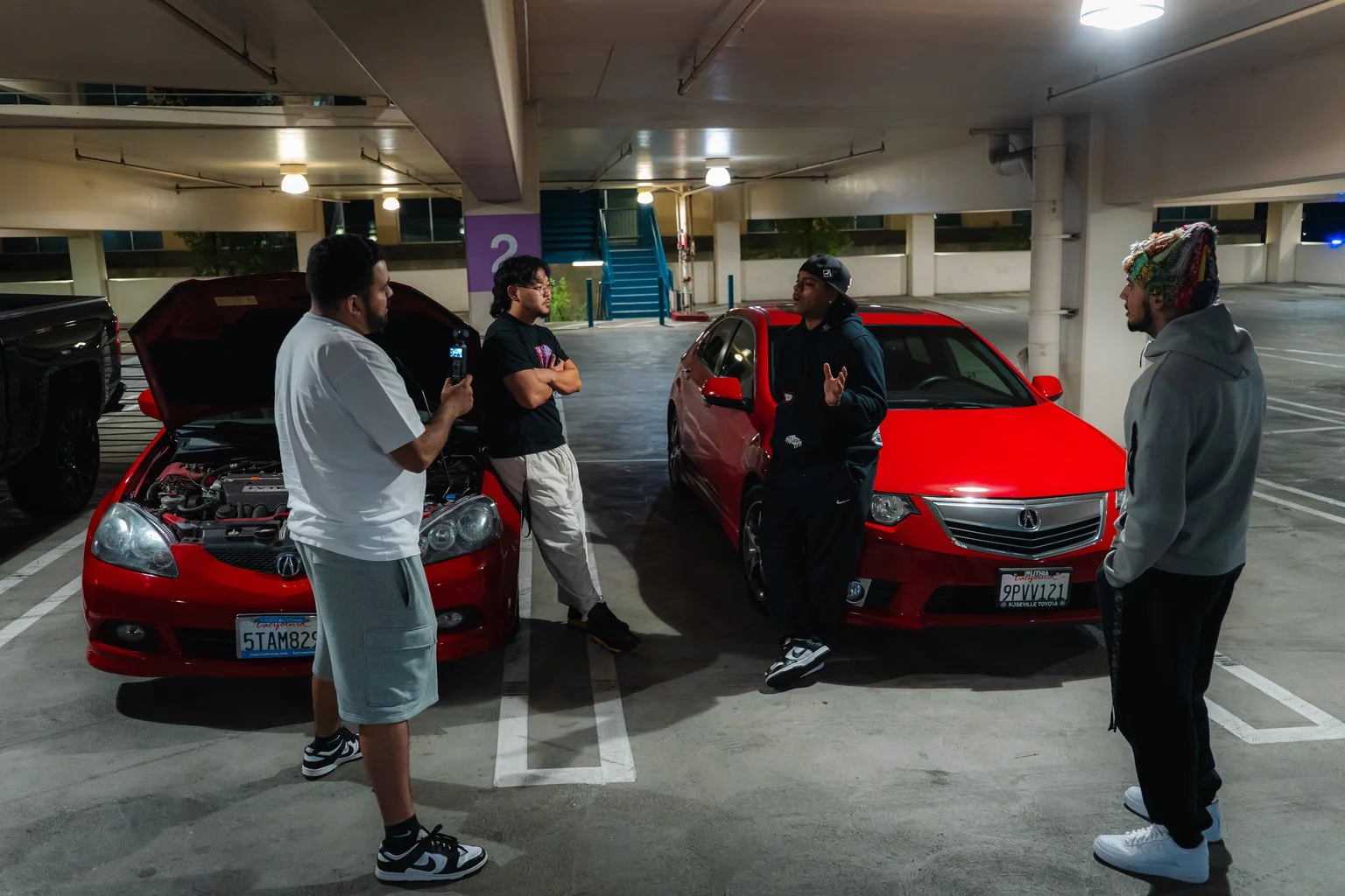 Five young men in casual clothing are standing in a parking garage, engaging in conversation. Two red cars are parked nearby, one with its hood open. The garage is well-lit with some empty parking spaces around.