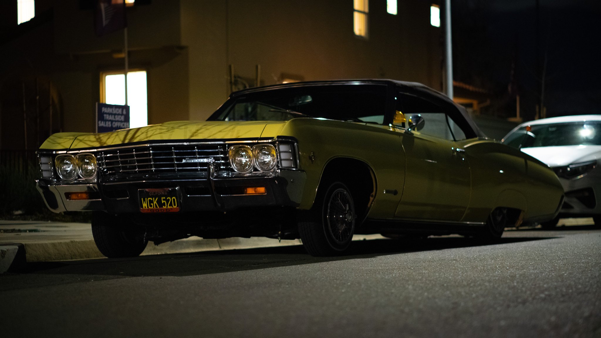 A vintage yellow and black car parked on the street at night, with a building with lit windows in the background.