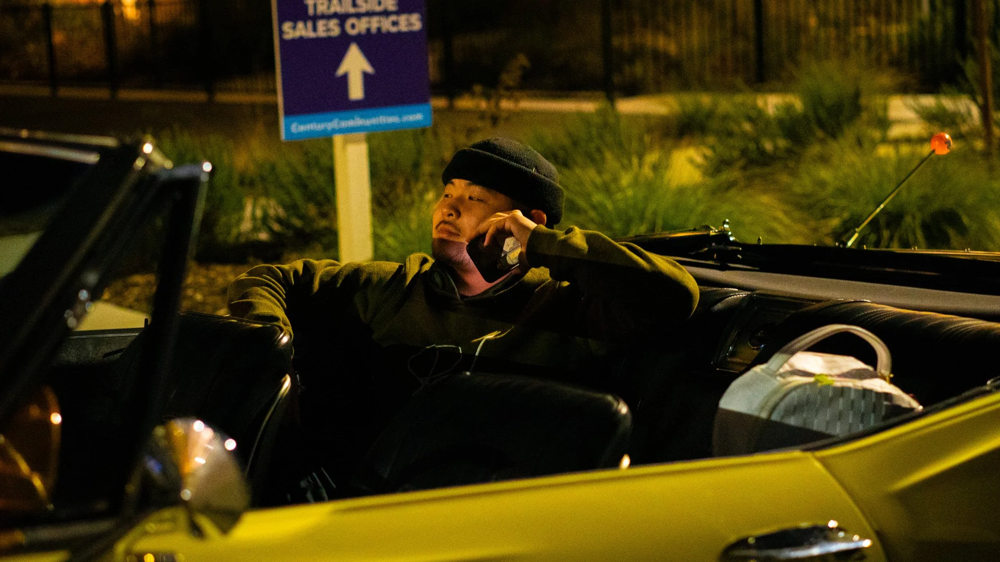Person sitting in a yellow convertible car talking on a cellphone at night, with a sign for sales offices in the background.