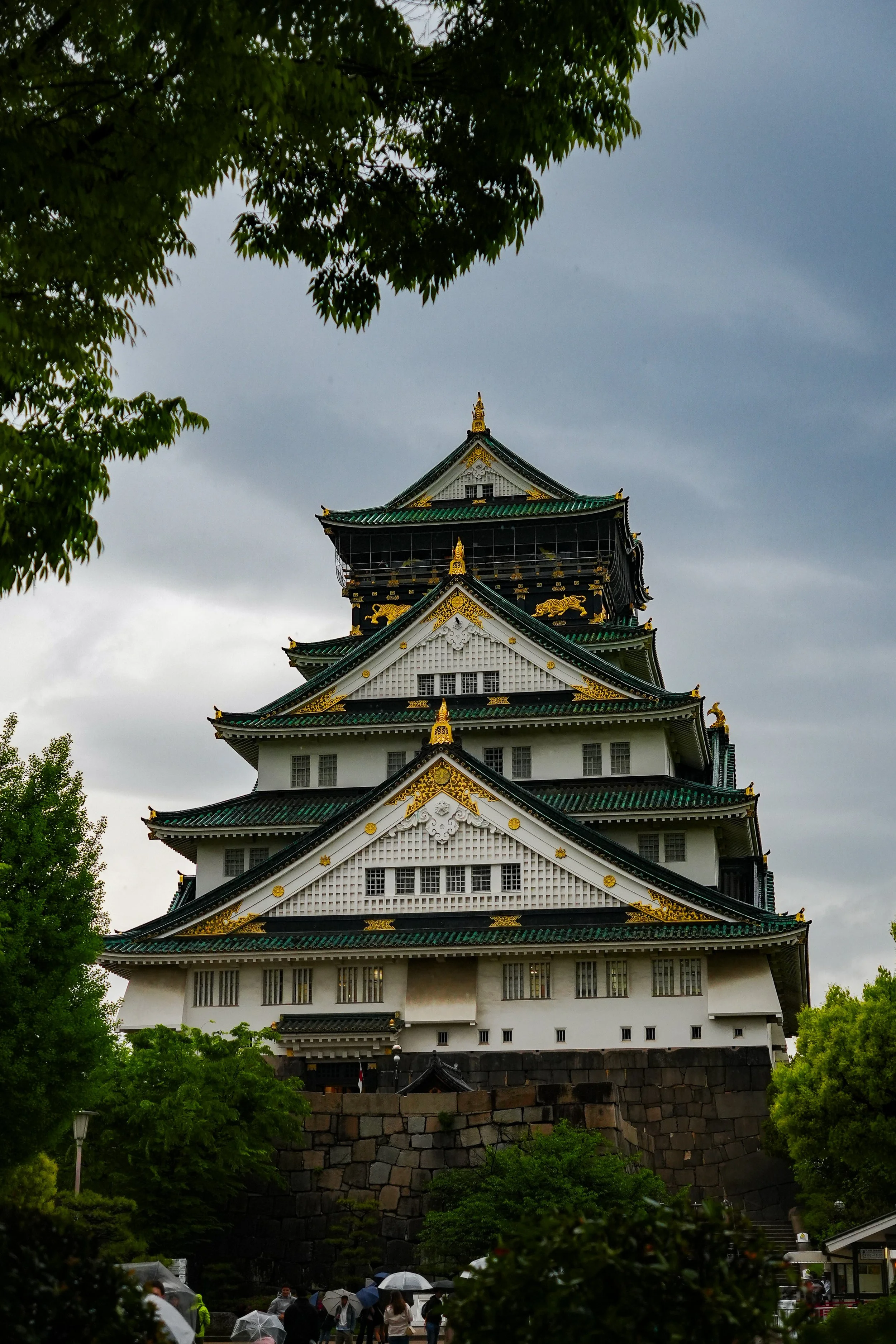 Traditional Japanese castle with multiple tiers, black and white walls, green roof tiles, and gold decorative elements, surrounded by green trees under a cloudy sky with people holding umbrellas in the foreground.