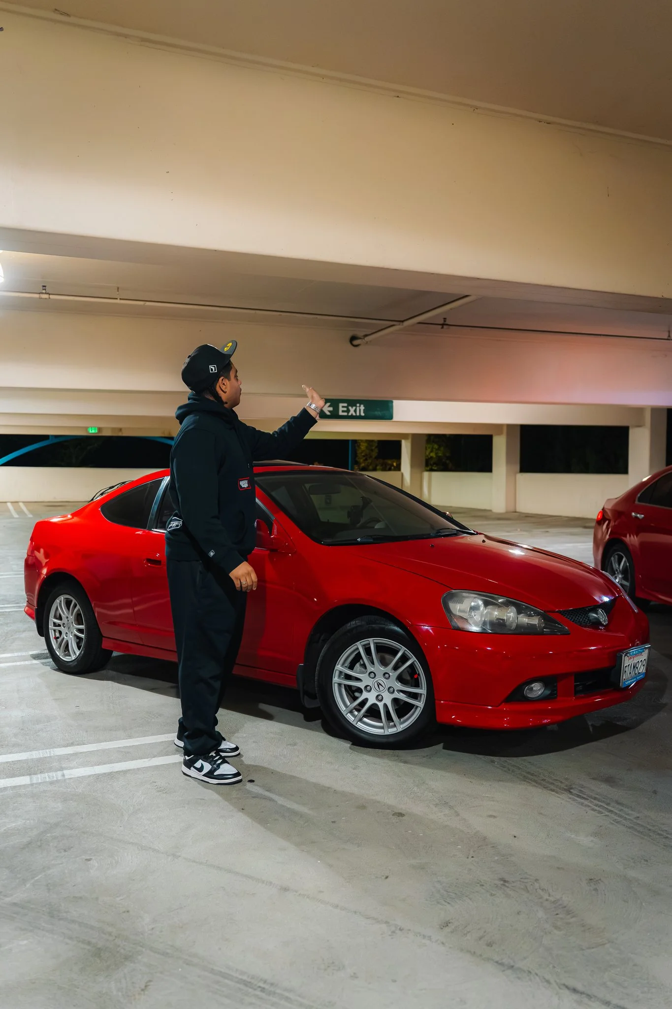 A man in a black hoodie and black pants, wearing a black cap and white sneakers, stands near a red car in an indoor parking garage, pointing towards the ceiling near an exit sign.