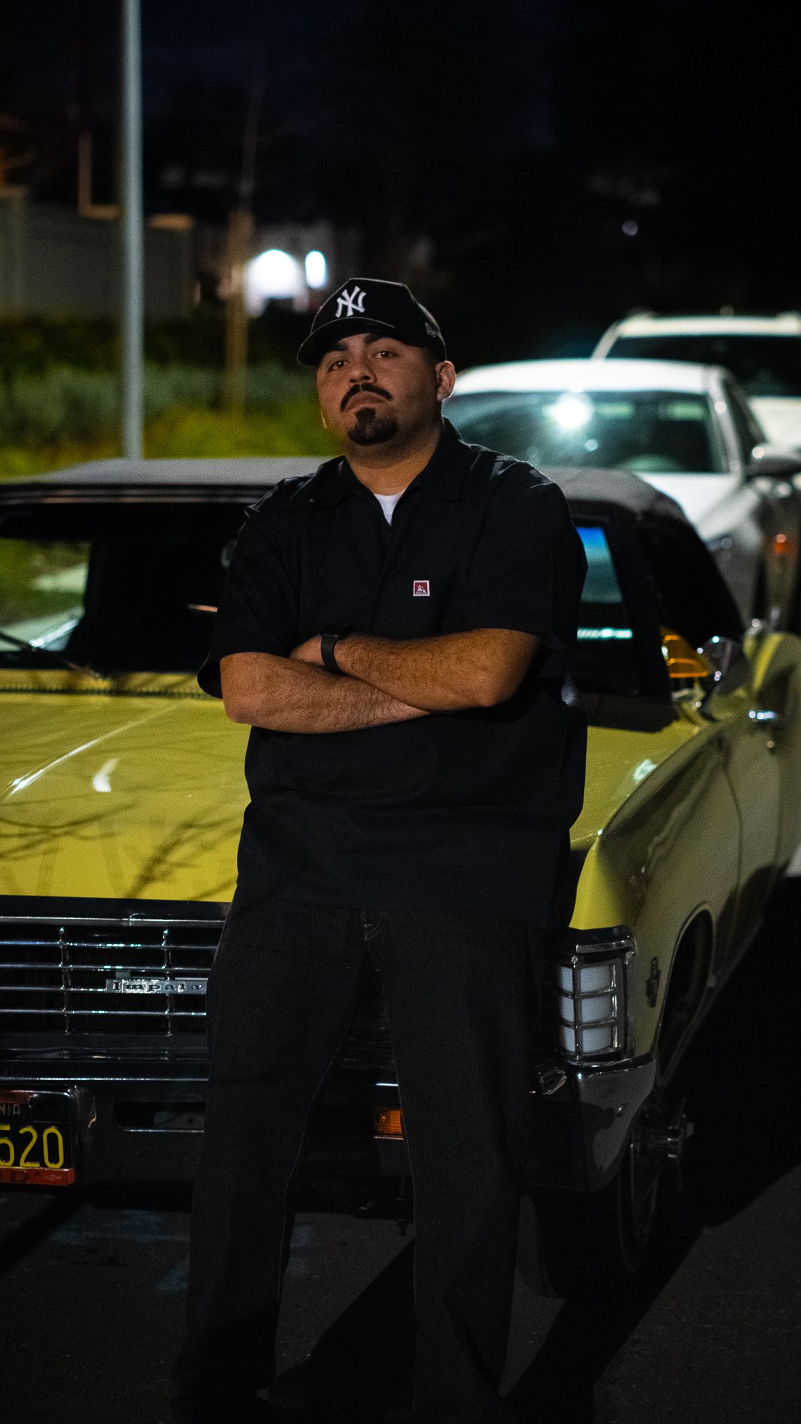 A man standing in front of a yellow classic car at night, with arms crossed, wearing a black NY cap and black shirt.