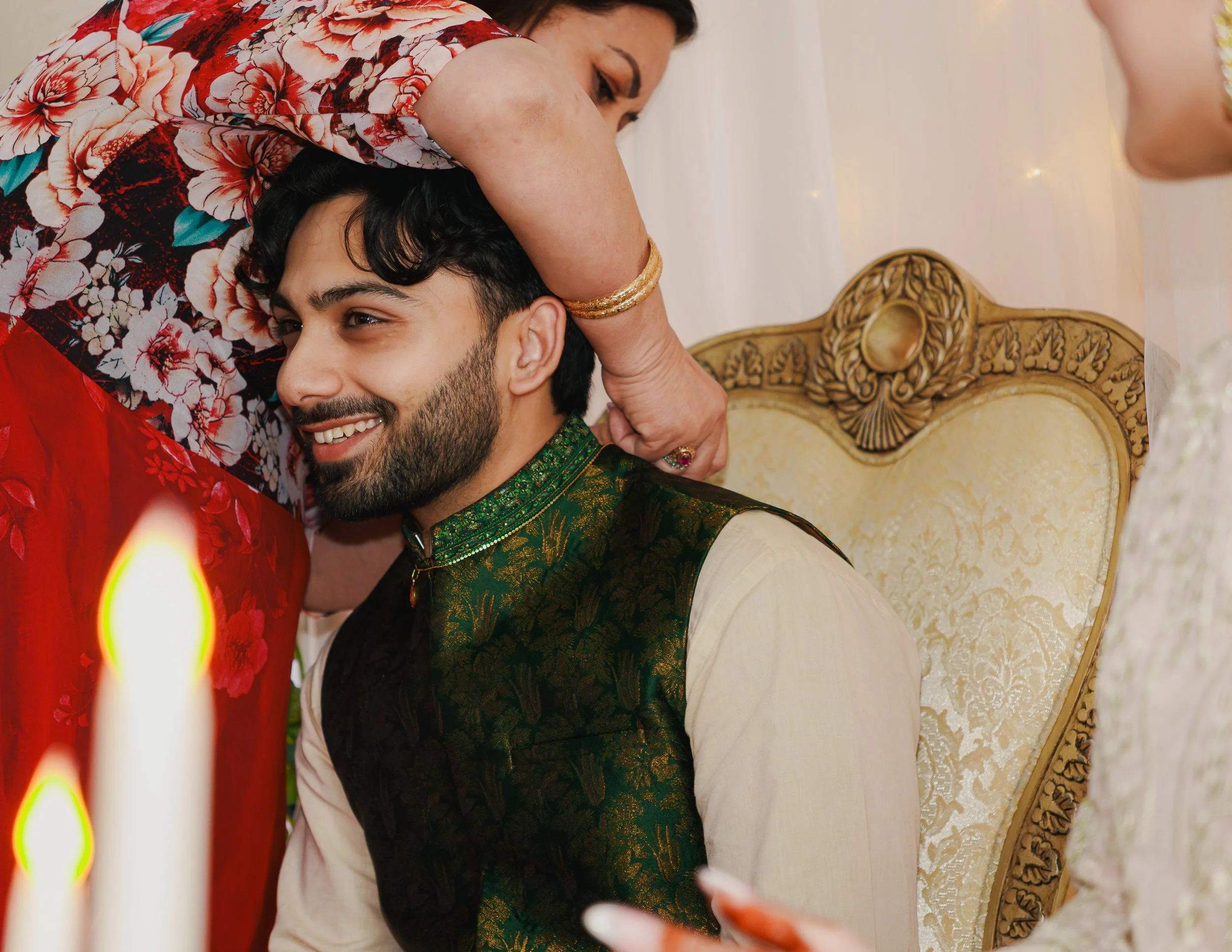 A woman kissing a young man on the forehead during a celebration, both dressed in traditional Indian attire, with ornate furniture and candles visible in the background.