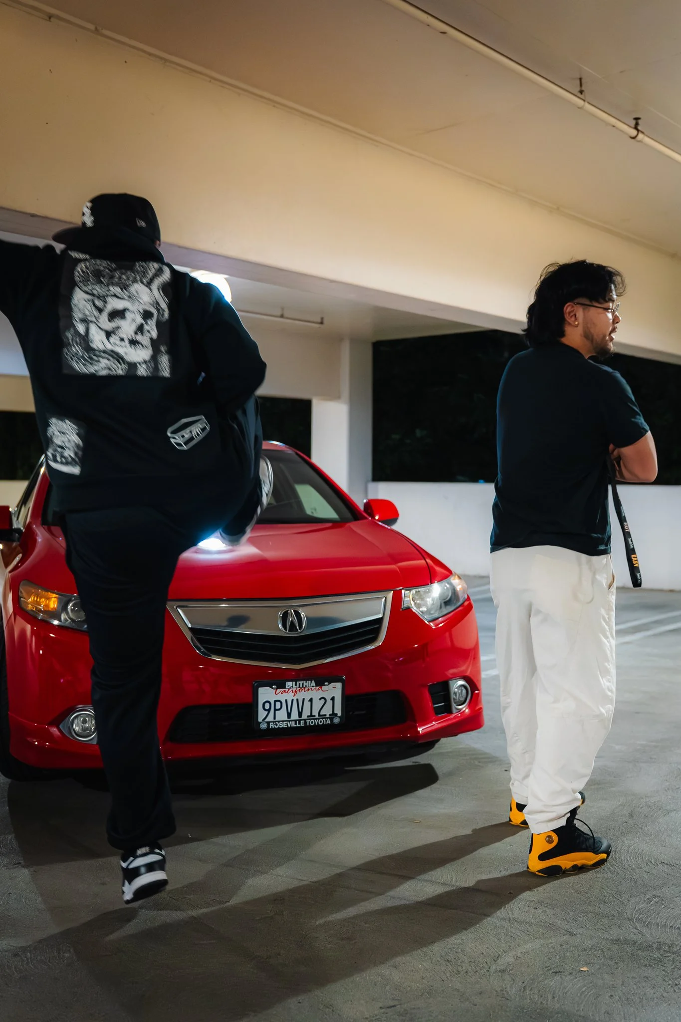 Two men are standing next to a red Acura car in a parking garage at night. The man on the left, wearing black clothing with a skull graphic on the back, has his arm extended and is facing away from the camera. The man on the right is wearing a black 