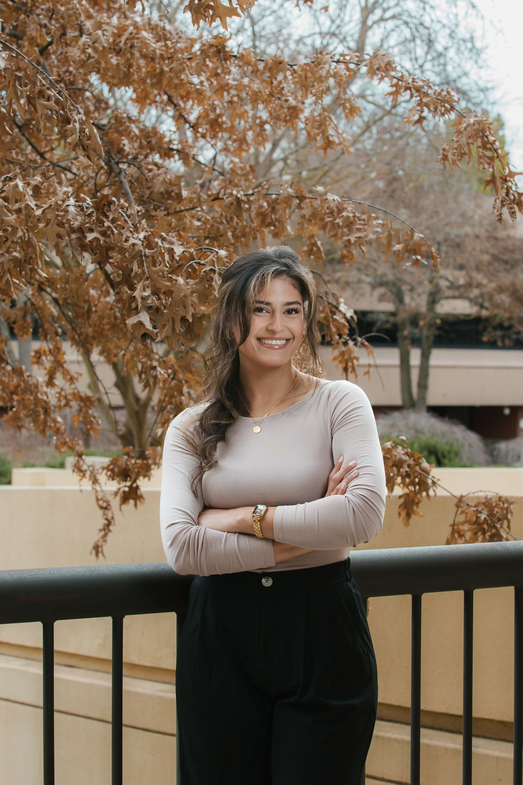 A smiling woman with long, wavy dark hair and light skin is standing outdoors, arms crossed, in front of a beige railing, wearing a beige long-sleeve shirt, black pants, a gold watch, and a necklace, with trees and autumn leaves in the background.
