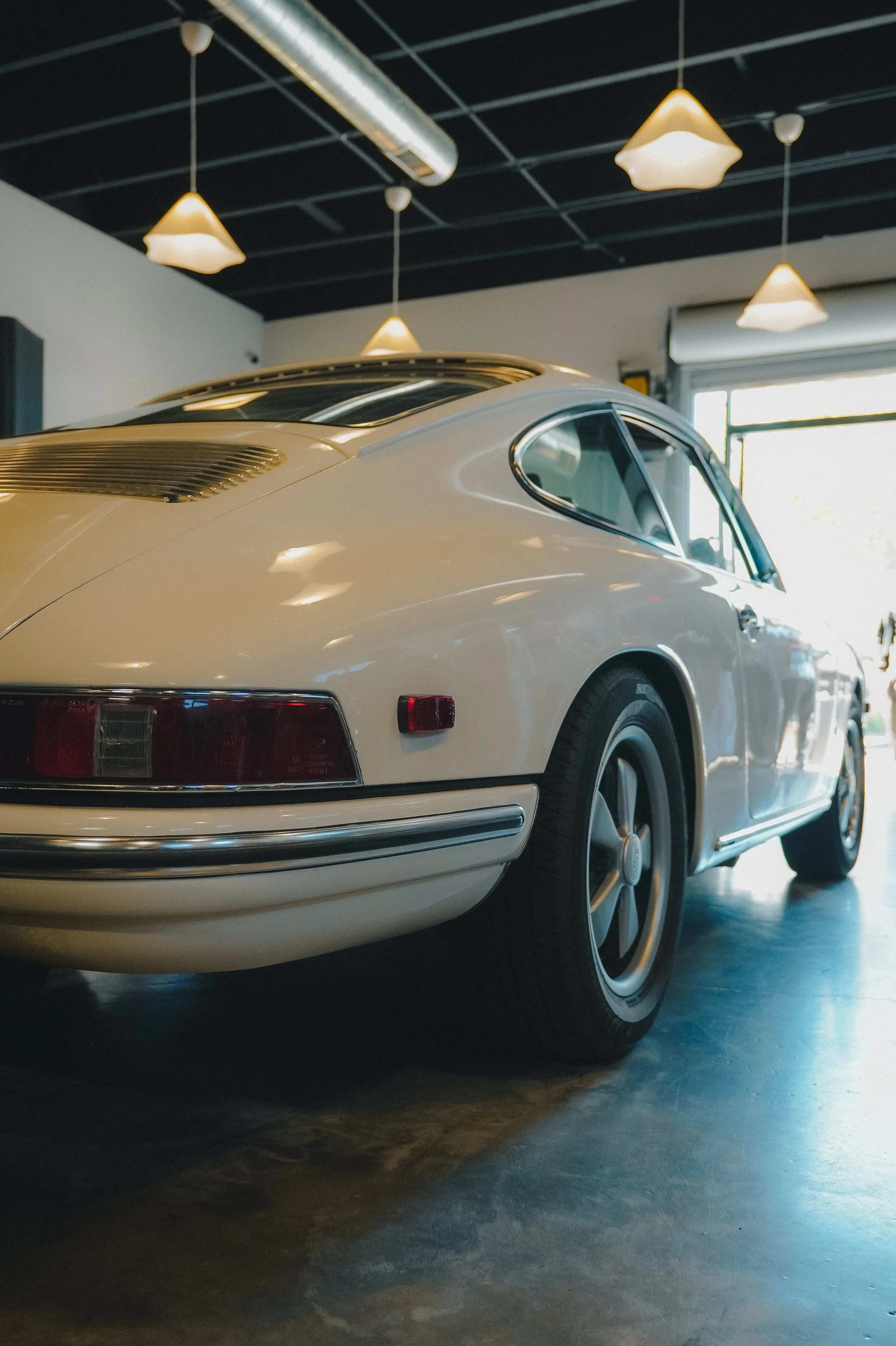 A white vintage sports car inside a garage, viewed from the rear right side, with a large window and ceiling lights in the background.