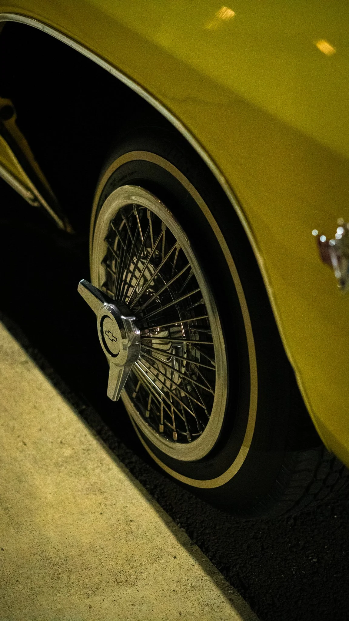 Close-up of a vintage yellow Chevrolet car wheel with wire spokes and Chevrolet emblem on the hubcap, parked on a light-colored concrete surface.