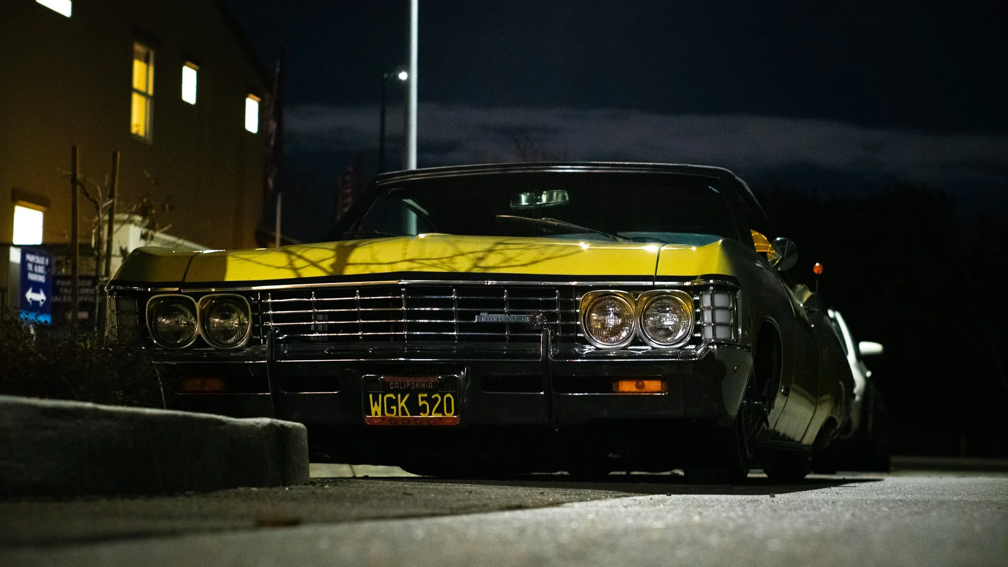 A vintage black and yellow Cadillac parked outside at night, with a California license plate reading 'WGK 520'. The car is partially illuminated by nearby streetlights, with a building in the background and dark, cloudy sky above.