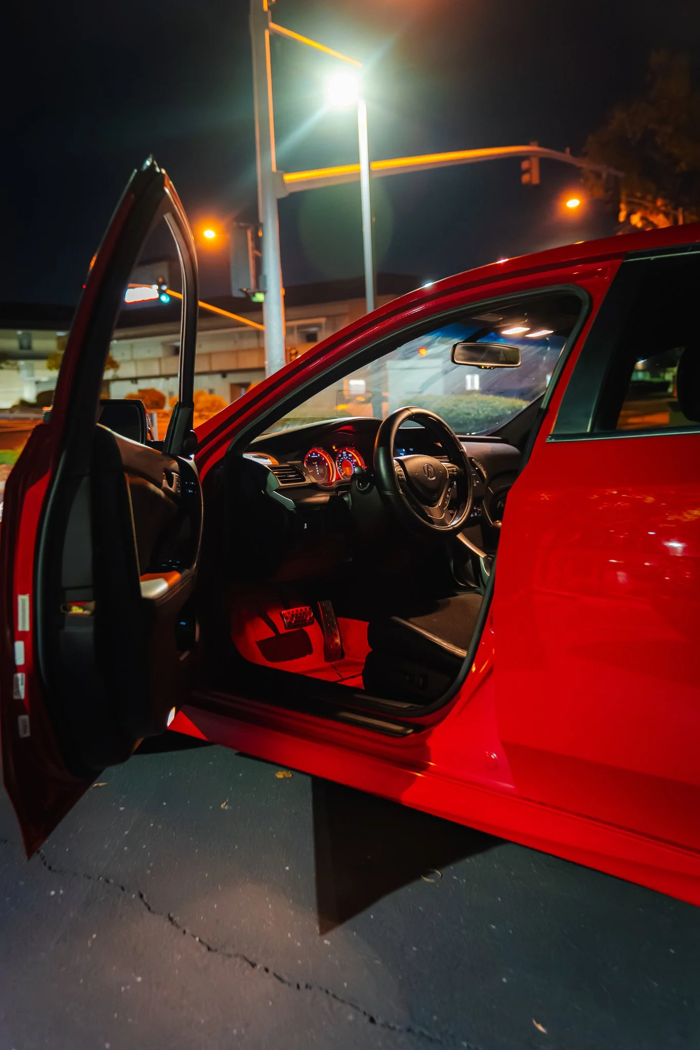 A red car with the driver's side door open, parked at night under streetlights, showing the interior dashboard and steering wheel.
