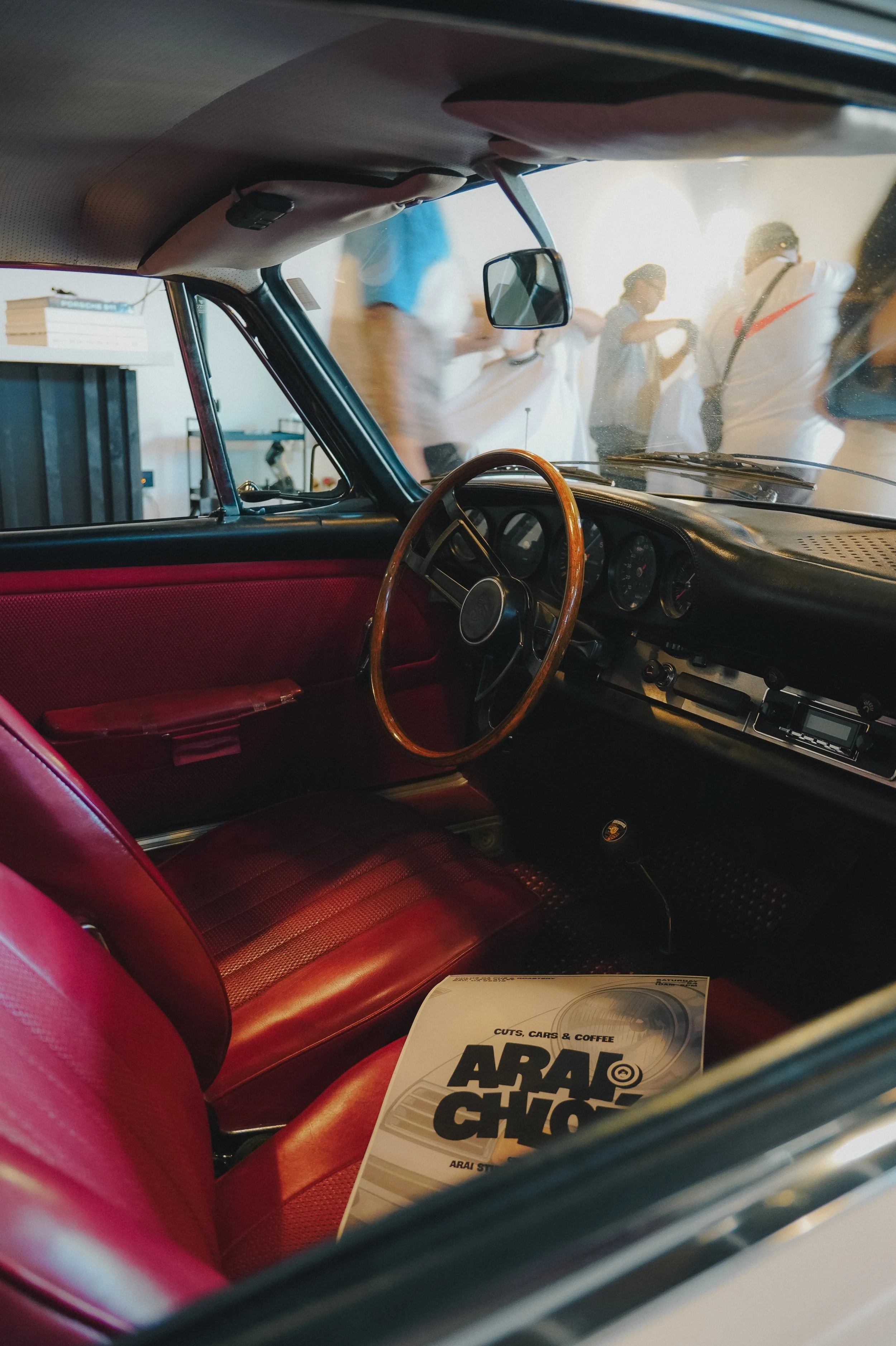 Inside a vintage car with a red leather seat, viewed through the window, with a brochure titled 'ARAI' on the seat, and people standing outside, some wearing sunglasses and white shirts.