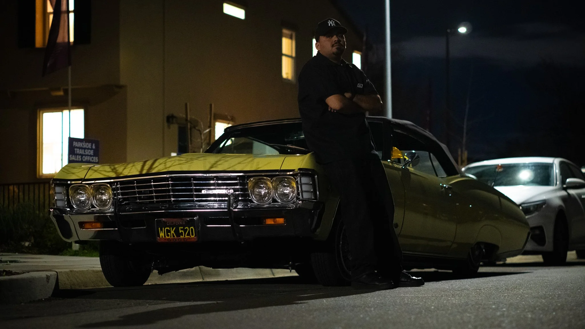 A man stands with arms crossed next to a vintage yellow car at night in a parking lot.