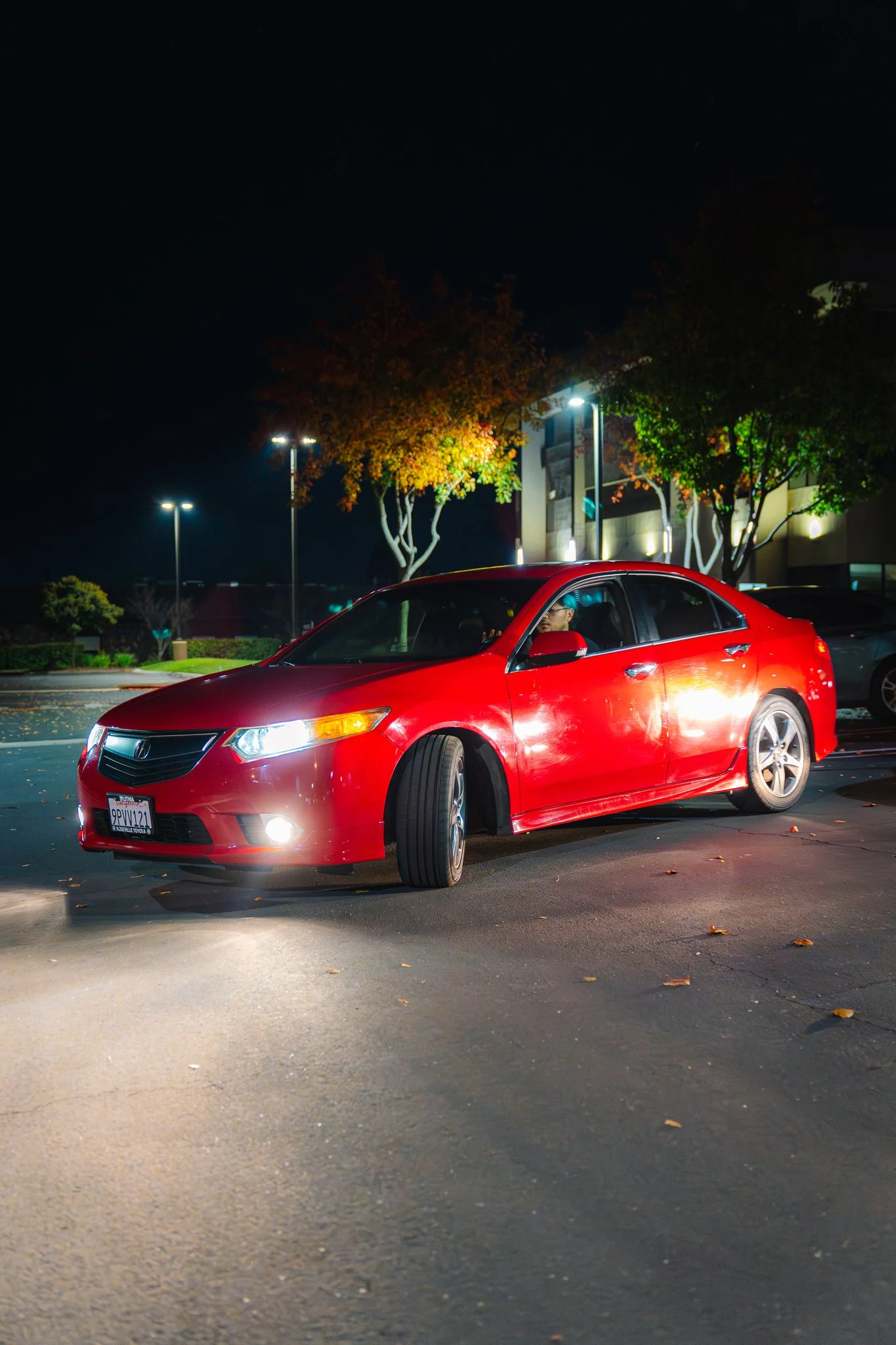 A red sedan car with its headlights on, parked in a illuminated parking lot at night. Trees and building lights are visible in the background.