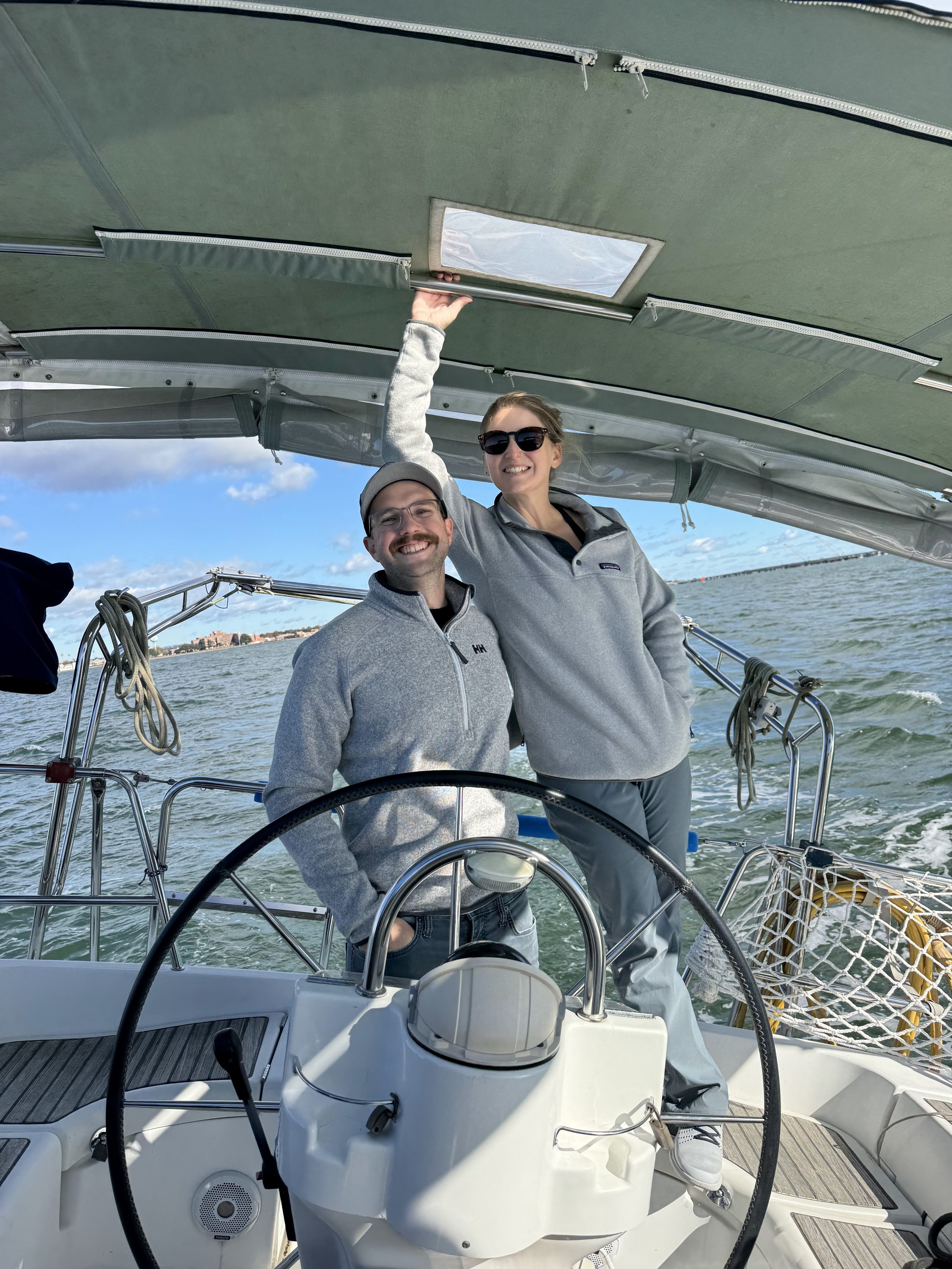 A man and woman standing on a boat, smiling, with the woman reaching up to open a hatch in the boat's canopy. The boat is on water with a distant shoreline visible.