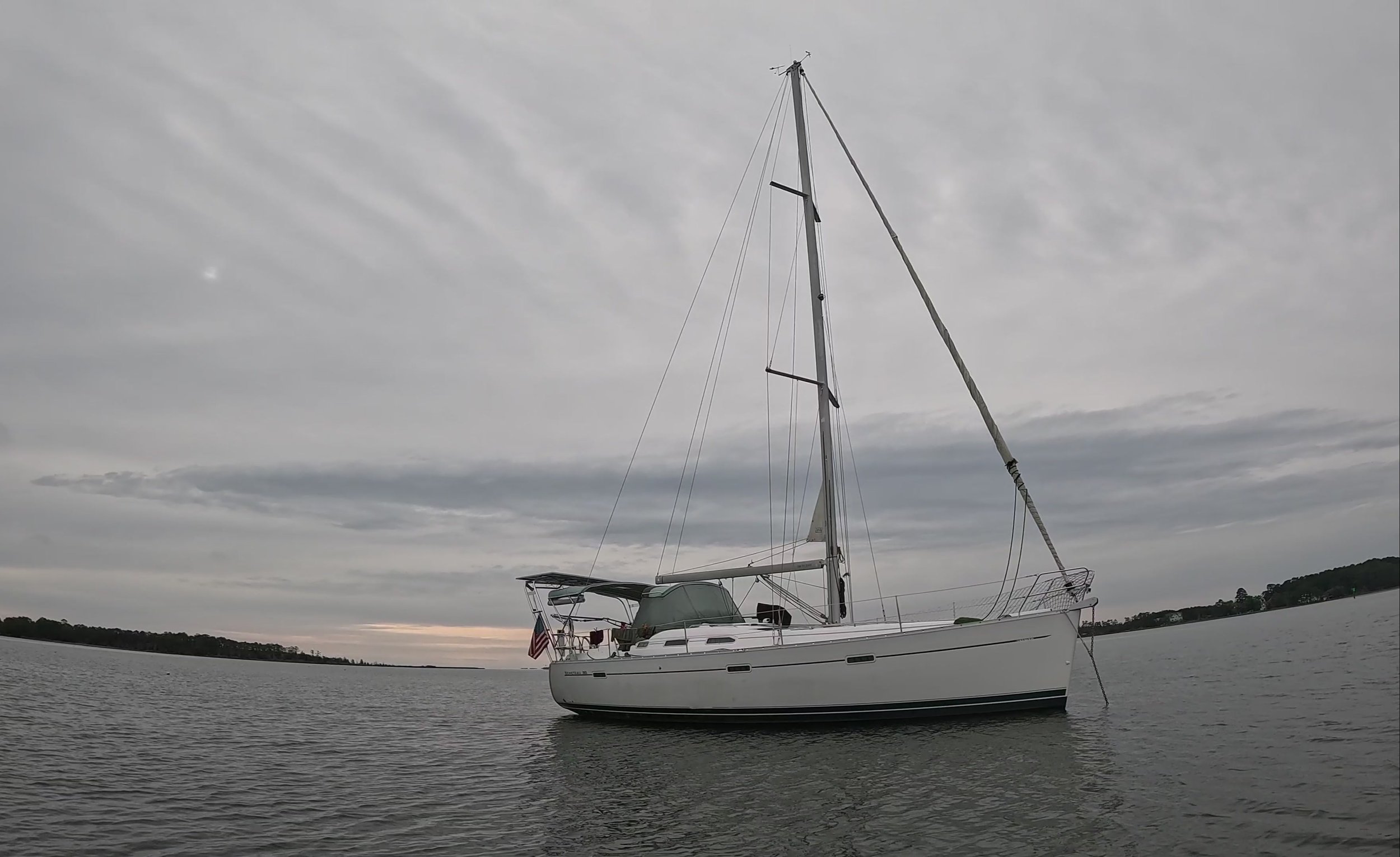 A white sailboat anchored on calm water under an overcast sky with a distant shoreline.
