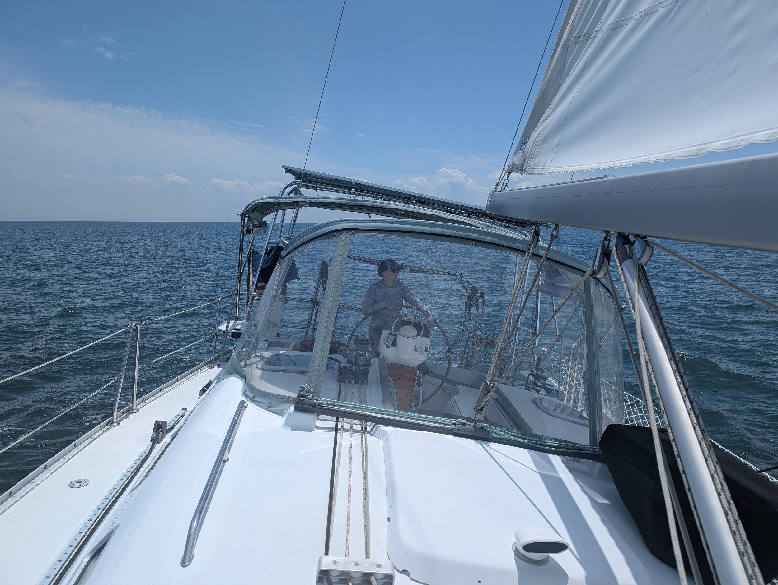 A person standing on a sailboat at sea, steering the wheel, with blue sky and ocean in the background.