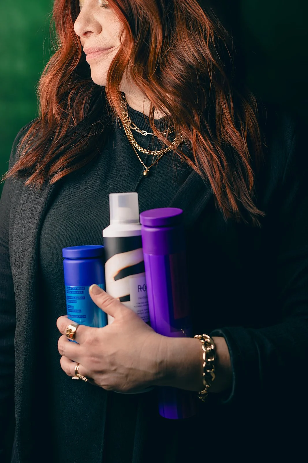 Woman with red hair holding three hair product bottles, wearing gold jewelry and a black outfit against a green background.