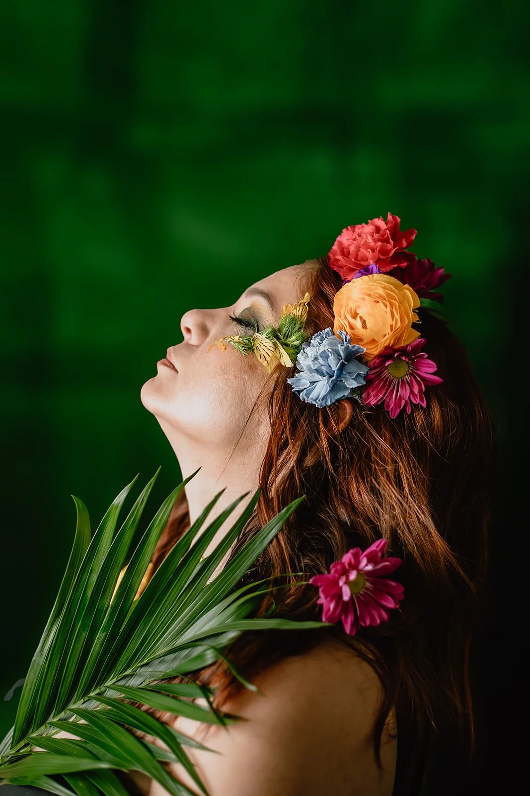 Woman with flowers in her hair and green leaves against a dark green background.
