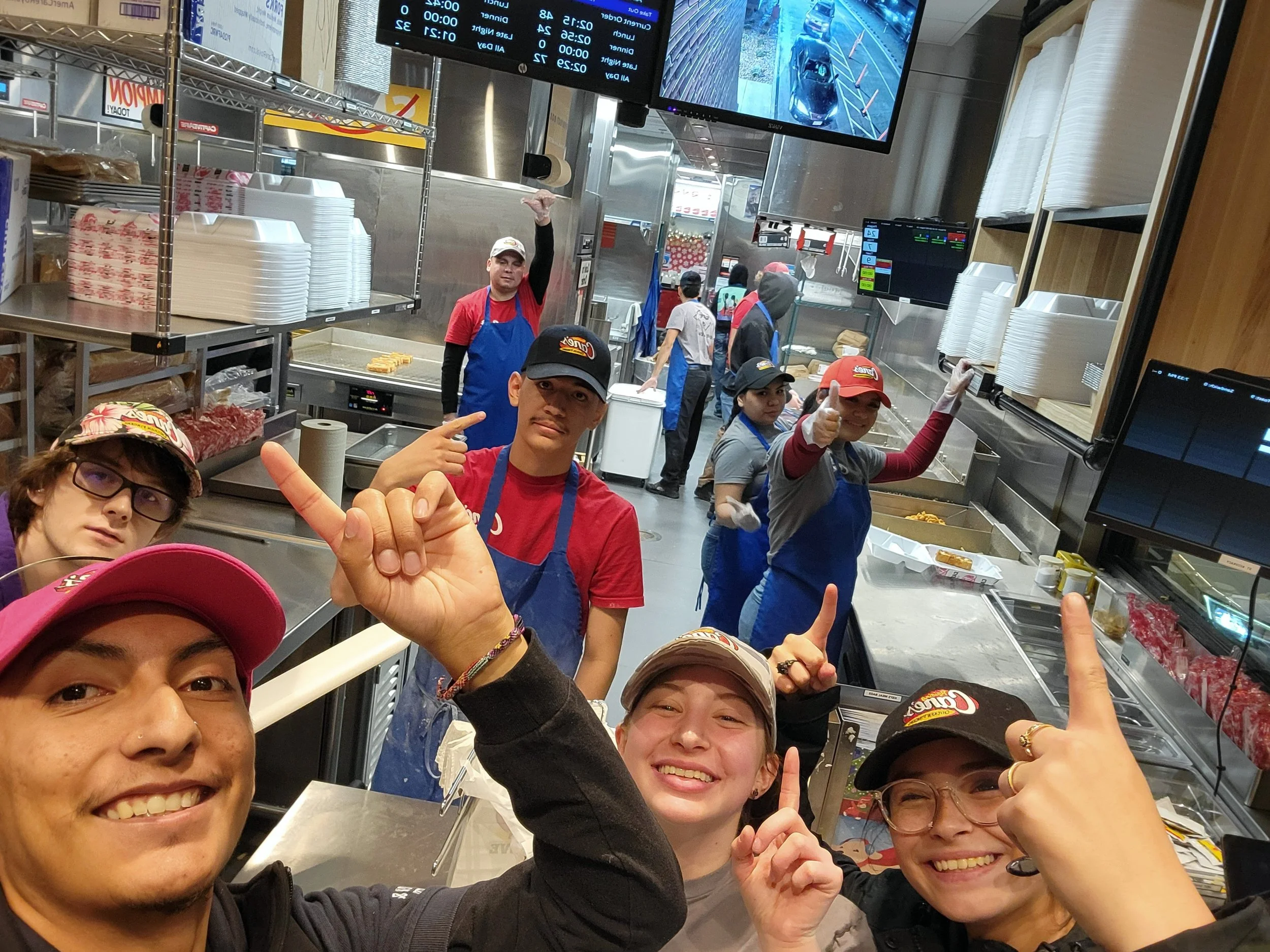 Gabriel and co-workers in a fast-food kitchen, smiling and pointing upwards, with two large screens displaying order information and camera footage behind them.