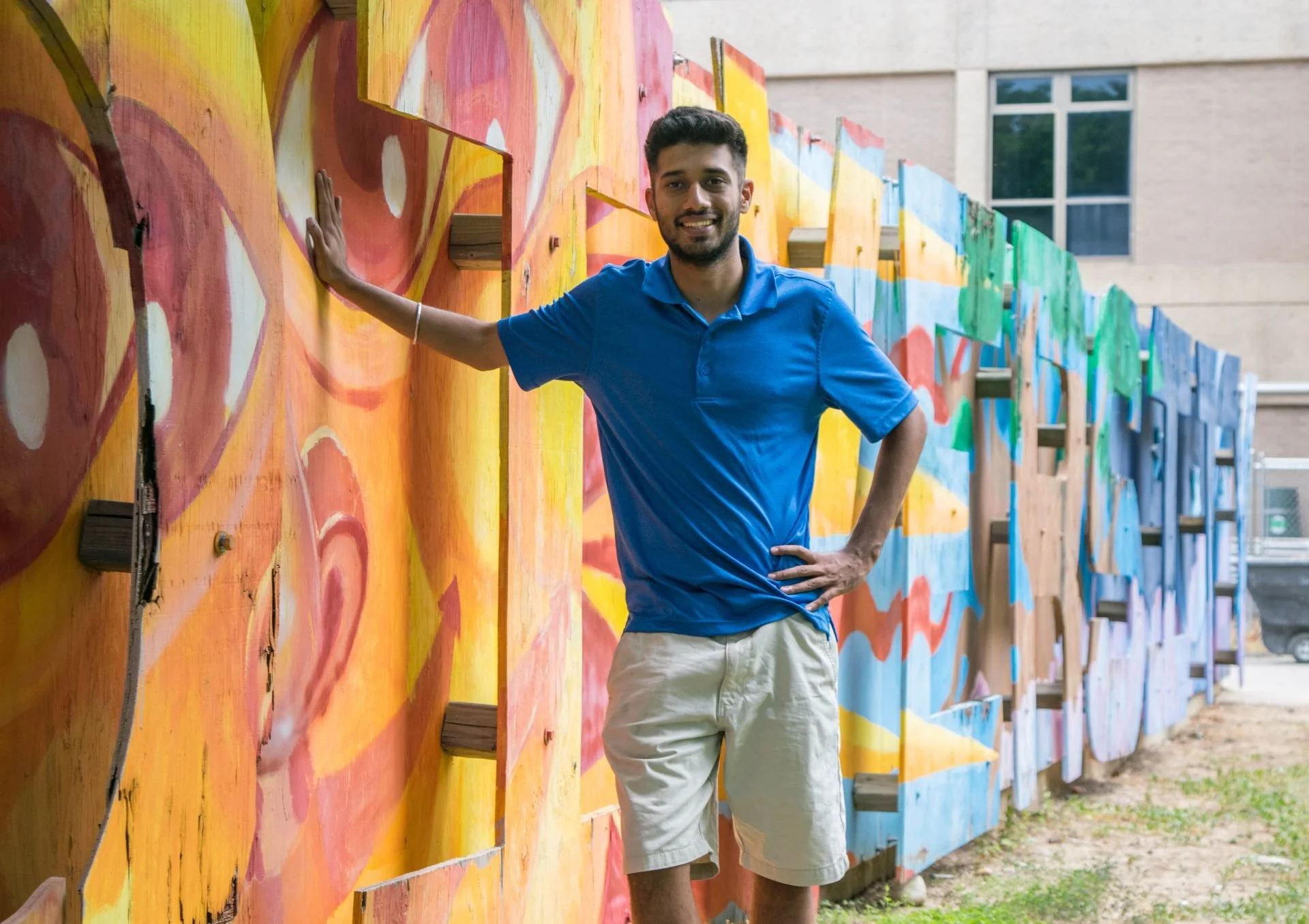 A young man standing outdoors next to a colorful, painted wooden fence, smiling at the camera, wearing a blue polo shirt and beige shorts.