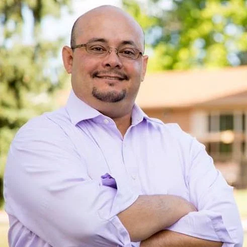 A man with a shaved head, glasses, and a goatee, smiling with arms crossed, wearing a light purple button-up shirt outdoors with trees and a house in the background.