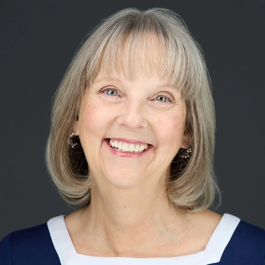 A smiling middle-aged woman with blue eyes, wearing earrings and a navy blue top with white accents, against a dark background.