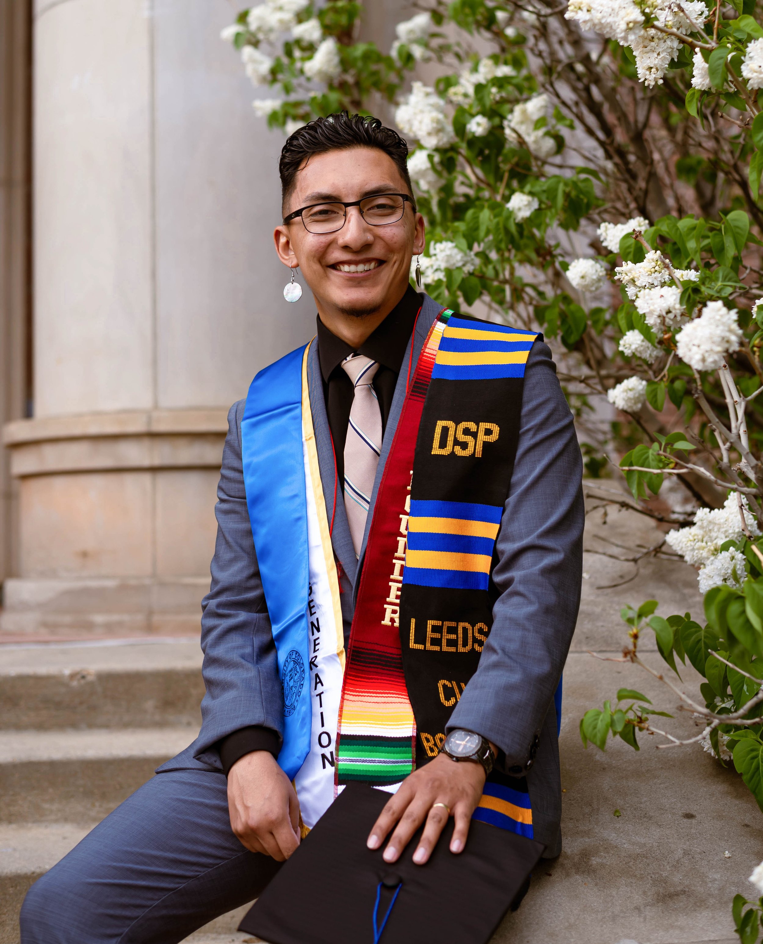 Gabriel Cervantes celebrating graduation, wearing a colorful stole and sash with various colors and patterns, sitting on steps near white flowers and greenery, smiling at the camera.