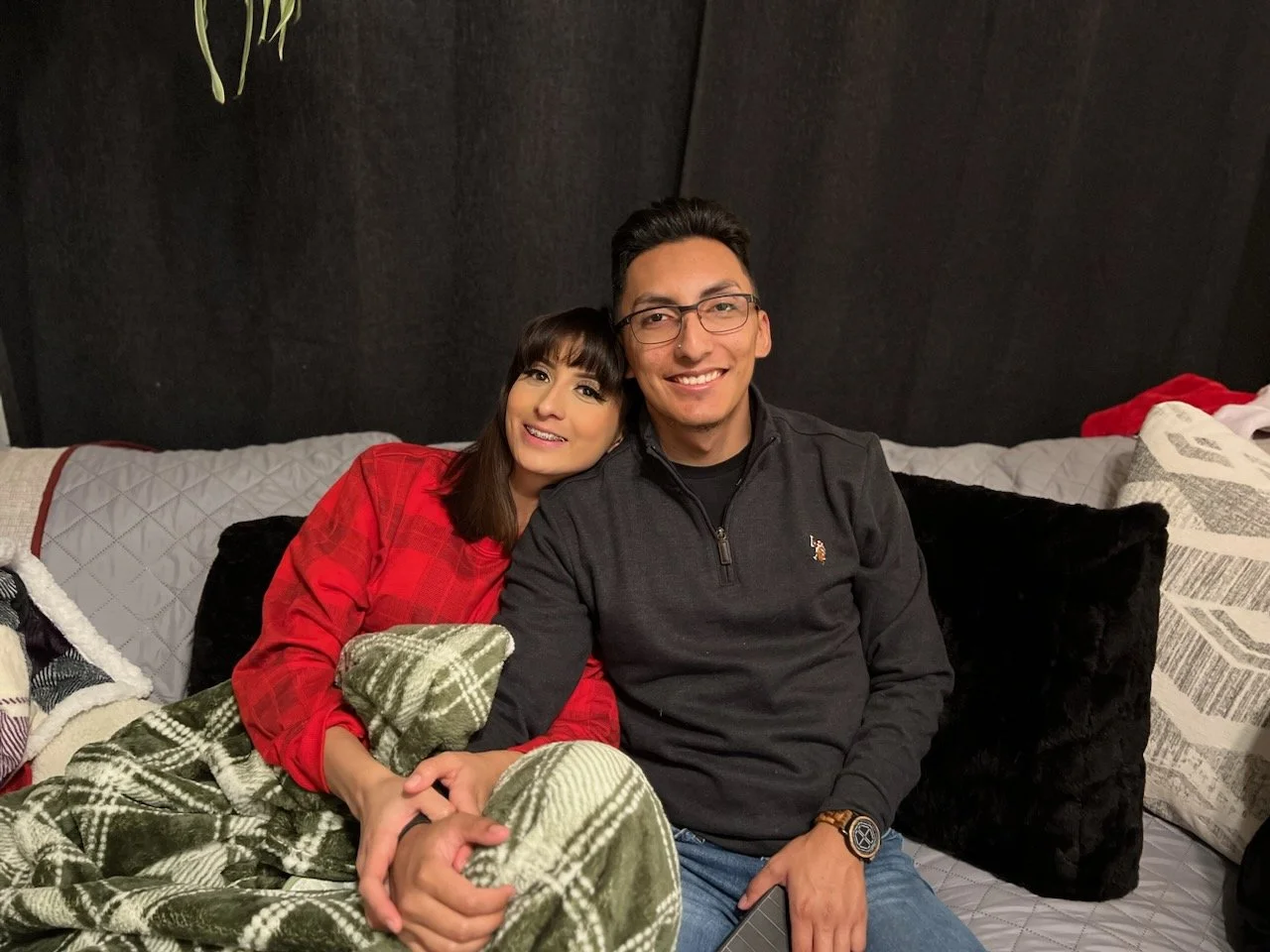 A smiling young man, Gabriel, and his wife sitting close on a couch, with the woman resting her head on the man's shoulder, surrounded by pillows and blankets.