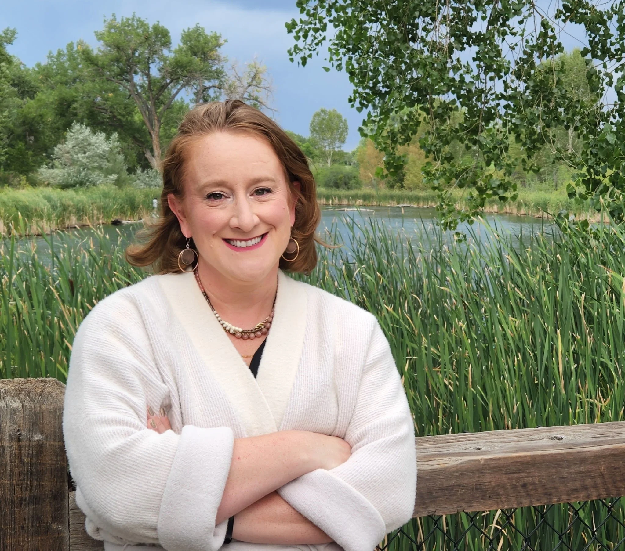 A smiling woman with brown hair, earrings, and a cream-colored sweater stands outdoors on a wooden bridge with arms crossed, in front of a body of water surrounded by green plants and trees, under a cloudy sky.