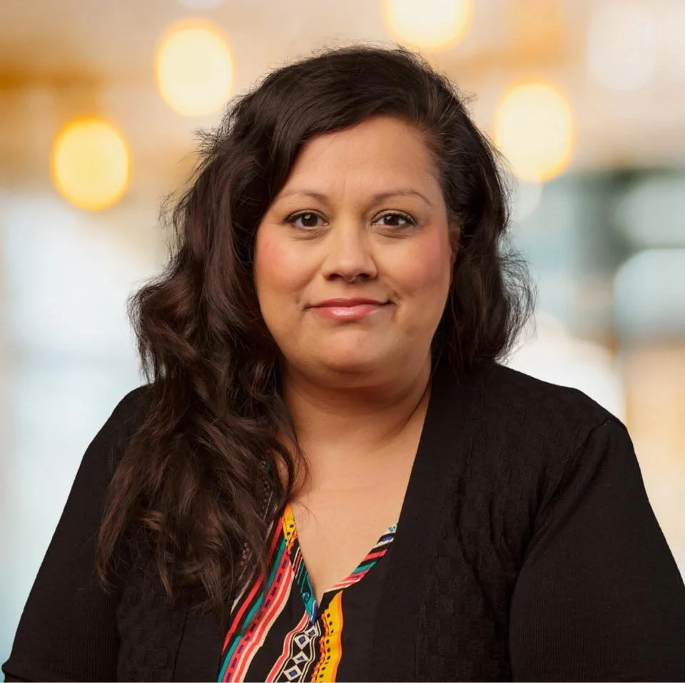 A woman with long, dark, wavy hair and light skin, wearing a black top with a colorful, patterned blouse underneath, smiling softly at the camera, with a blurred background of warm lights.
