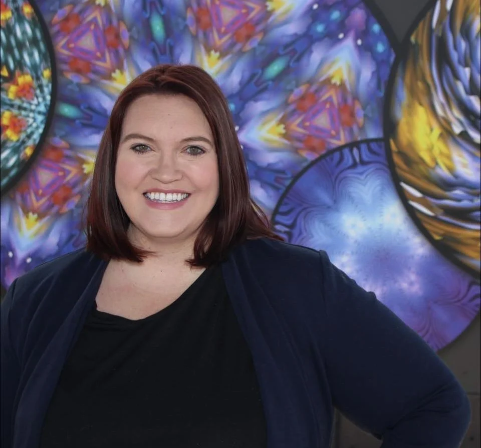 Smiling young woman with auburn hair standing in front of colorful abstract stained glass artwork.