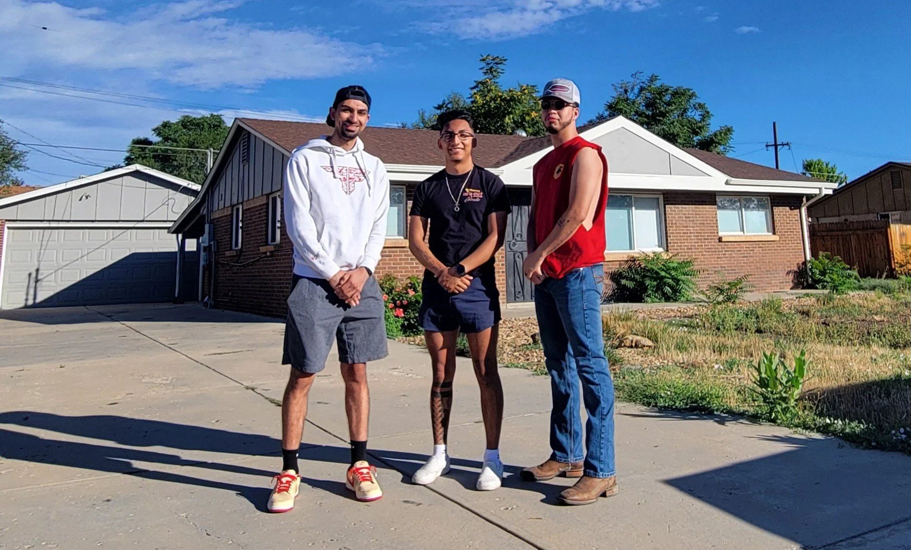 Gabriel and two friends standing on a driveway in front of a brick house with a white garage, smiling and posing for the photo on a sunny day.