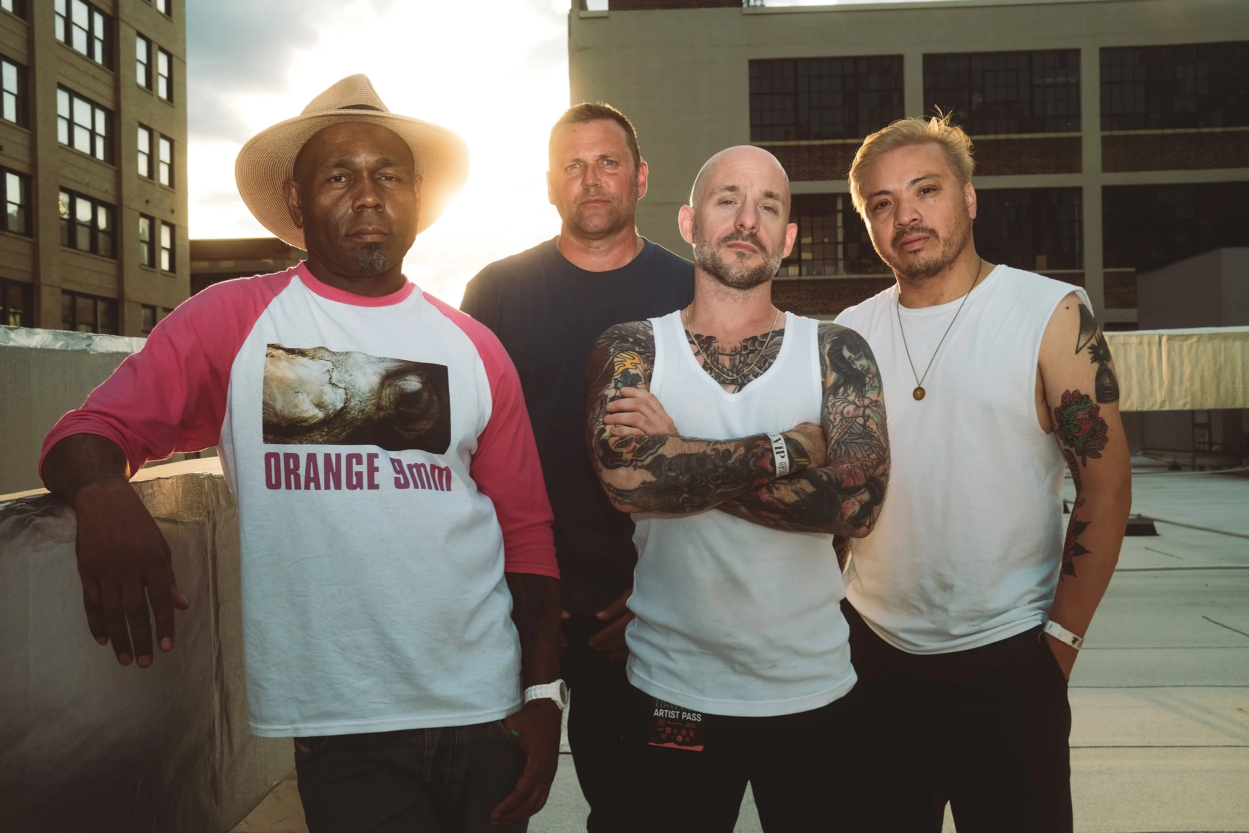 Group of five men standing on a rooftop in an urban setting during sunset, with tall buildings in the background. One man wears a wide-brimmed hat and a white shirt with pink sleeves and a graphic print. Another man has tattoos on his arms and is wearing a white sleeveless shirt. The other three men are dressed casually, with one in a black shirt, one in a black outfit, and one in a white tank top, all with various tattoos and accessories.