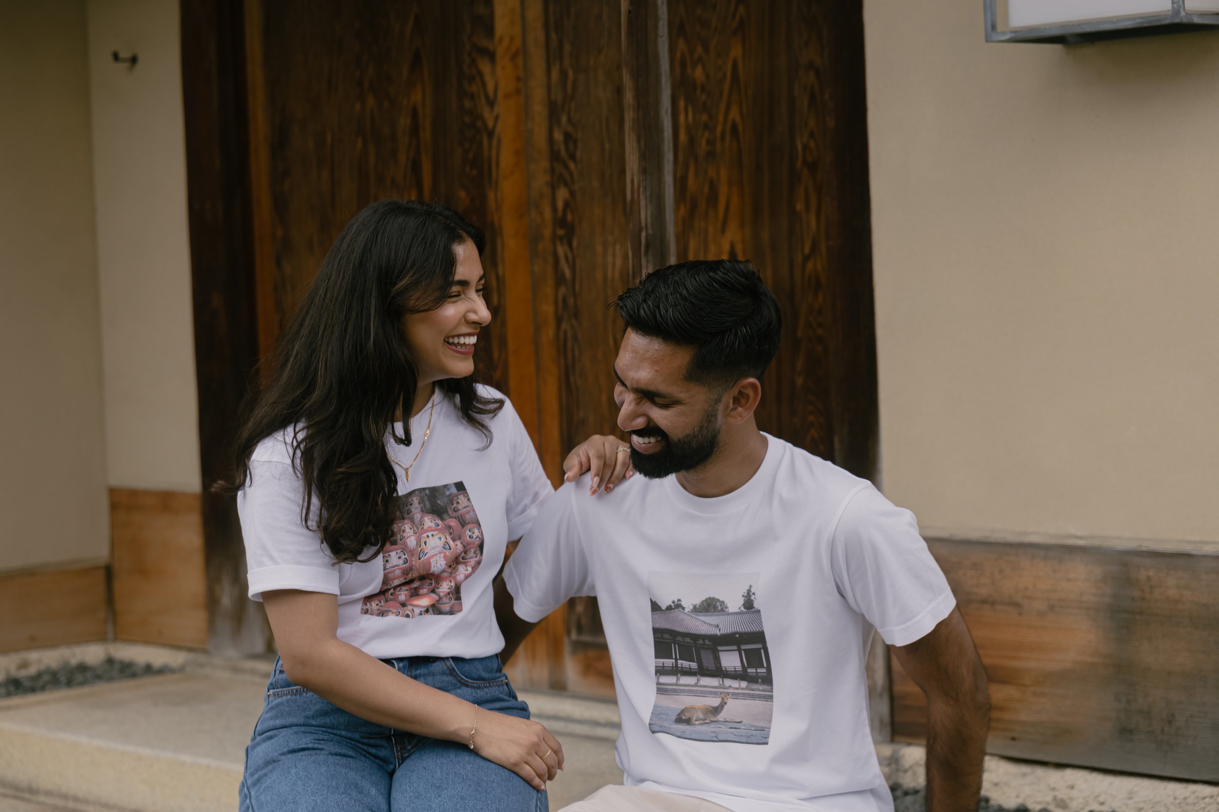 Two people, a woman and a man, are sitting close together, smiling and laughing. The woman has long dark hair and is wearing a white t-shirt with a colorful pattern, while the man has short dark hair, a beard, and is wearing a white t-shirt with a pr