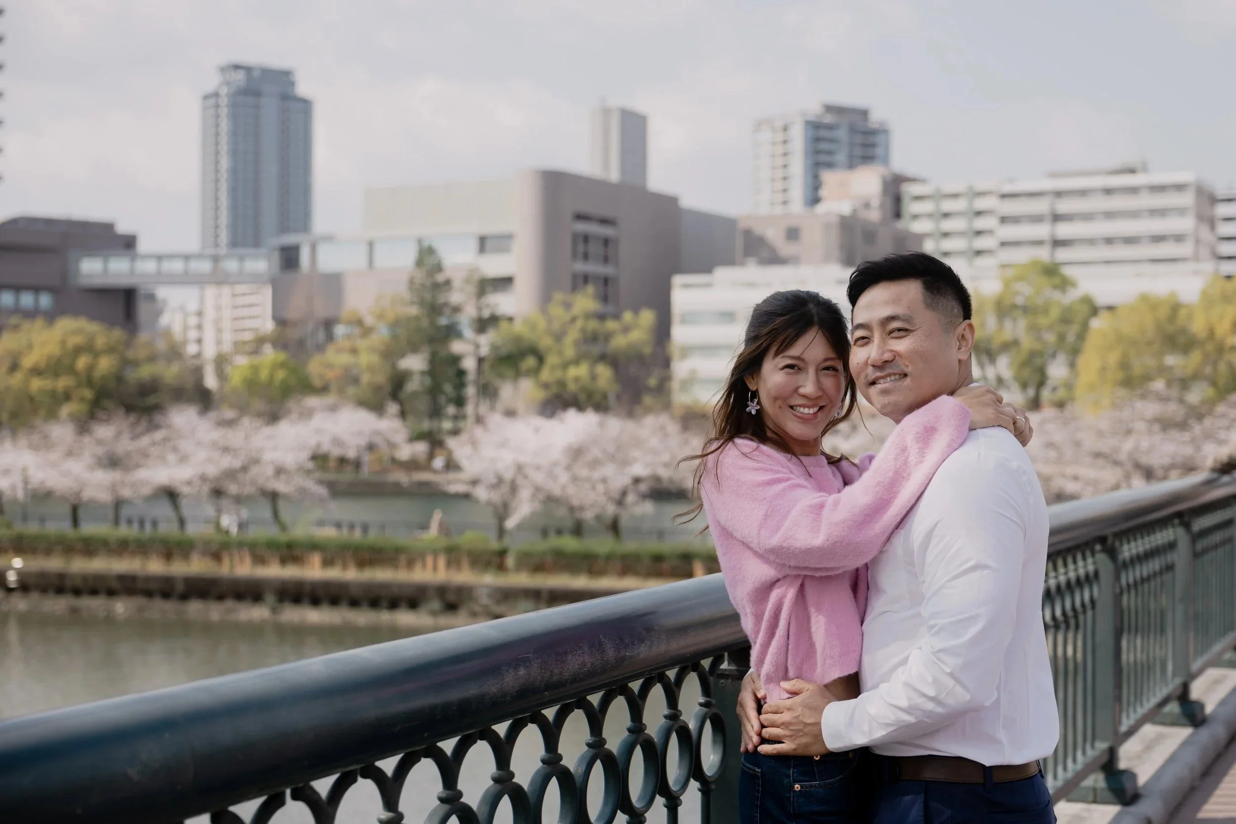 A couple hugging on a bridge with city buildings and cherry blossom trees in the background.